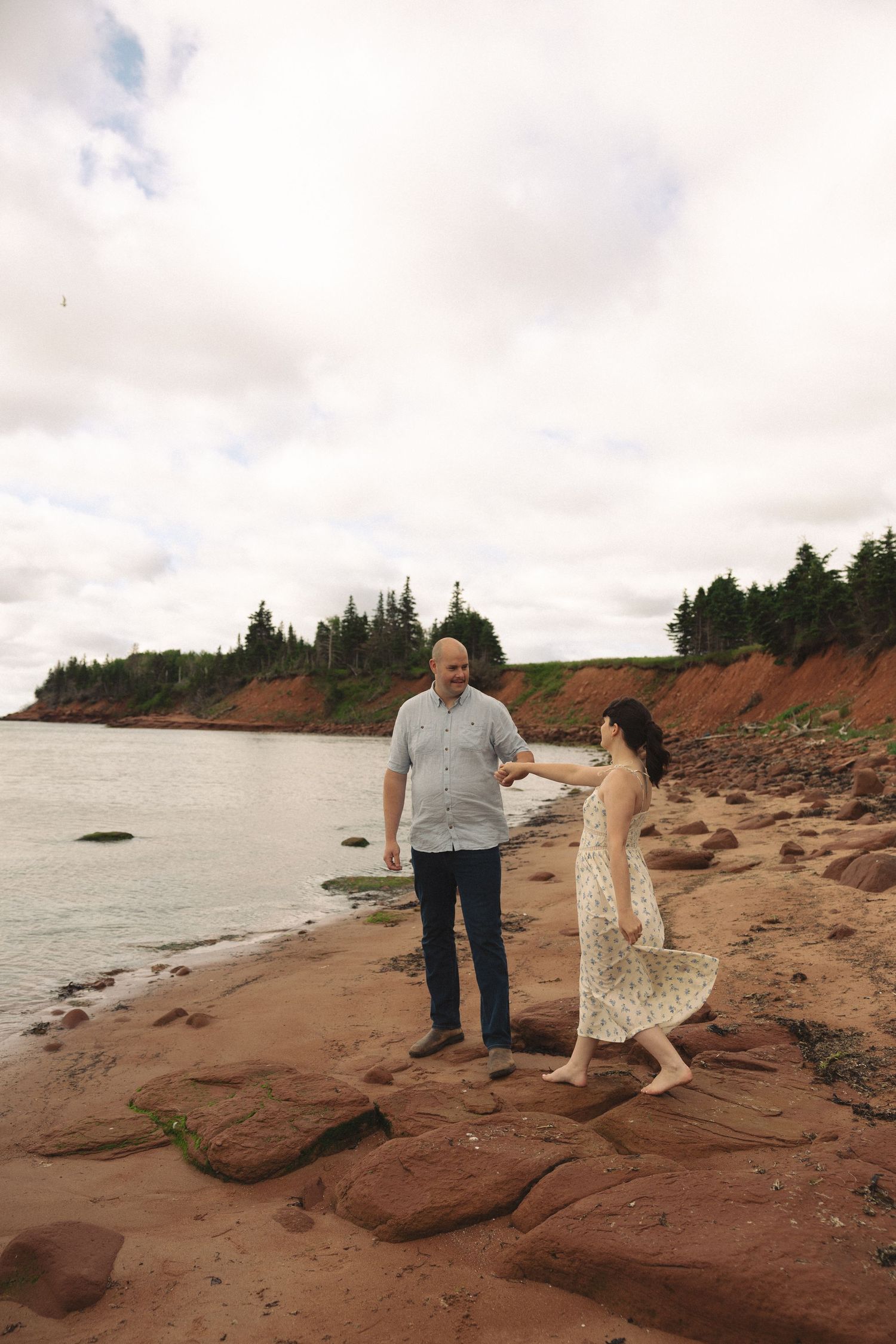 Couple holding hands on rocky red cliffs by the ocean with lush green trees in the background.