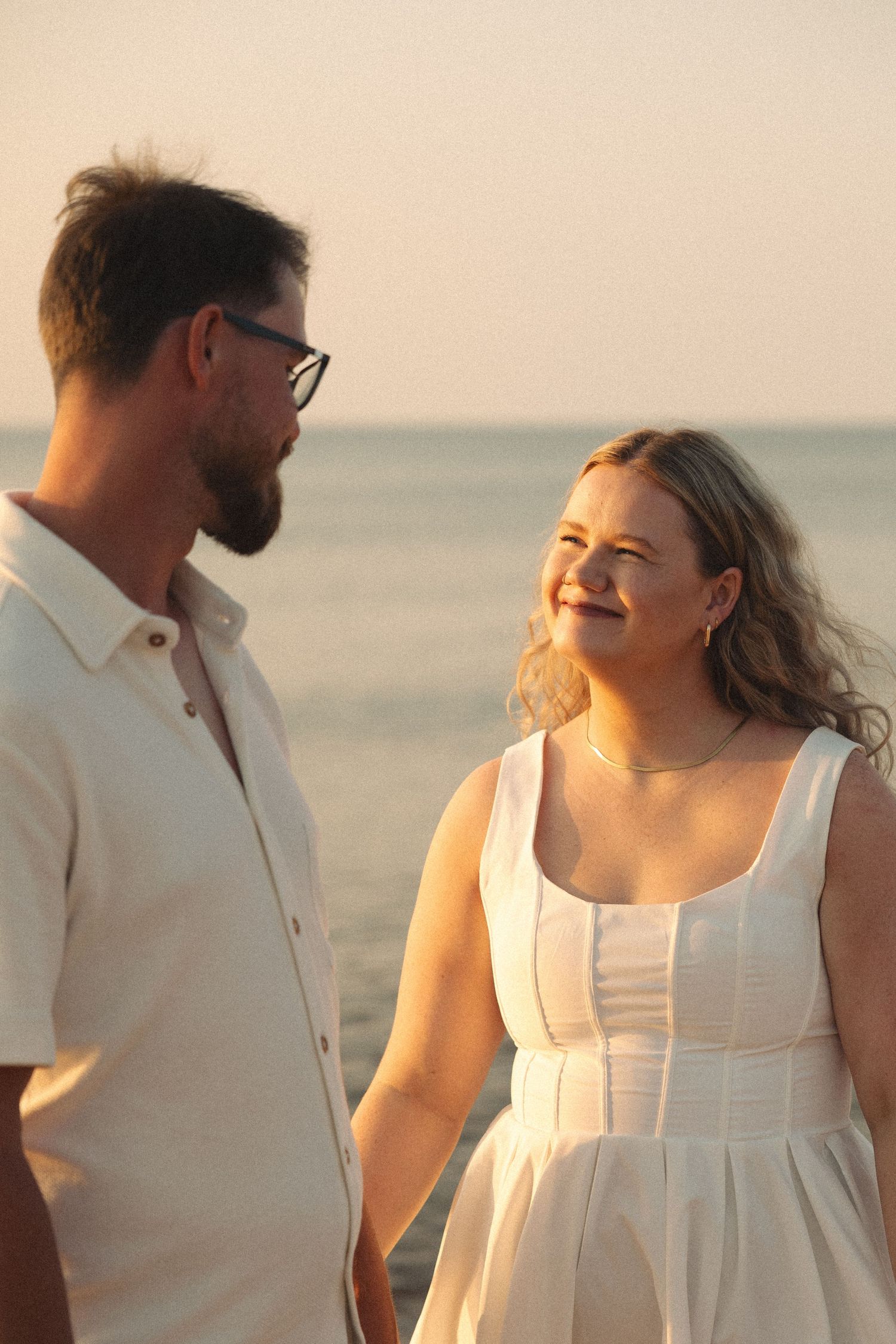 Smiling couple holding hands on beach at sunset, woman in white dress, man in light shirt.