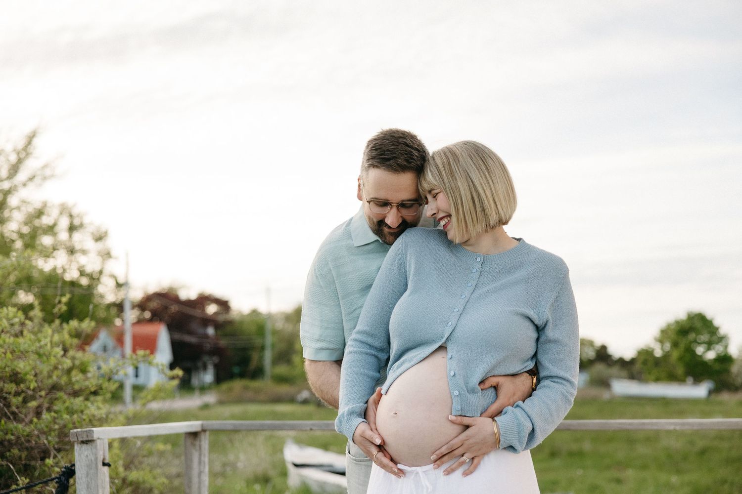 Happy couple embracing outdoors, man hugging pregnant woman in blue sweater while she holds her baby bump.