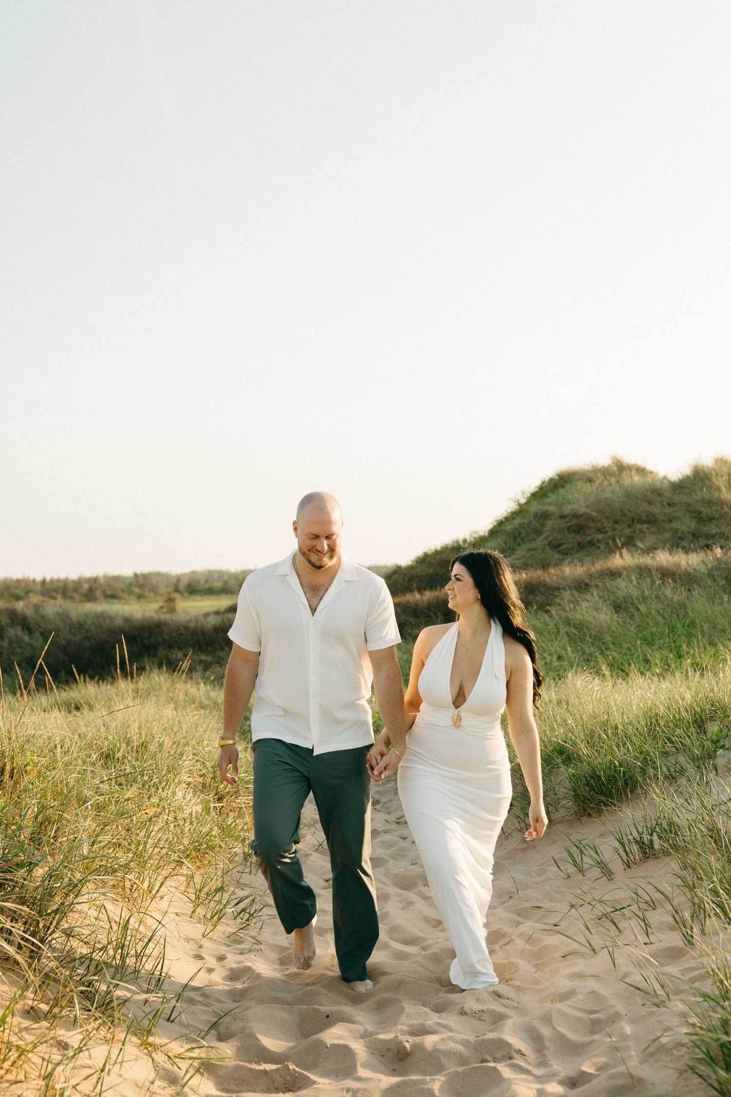 Couple holding hands walking barefoot on sandy beach path surrounded by green dunes at golden hour.