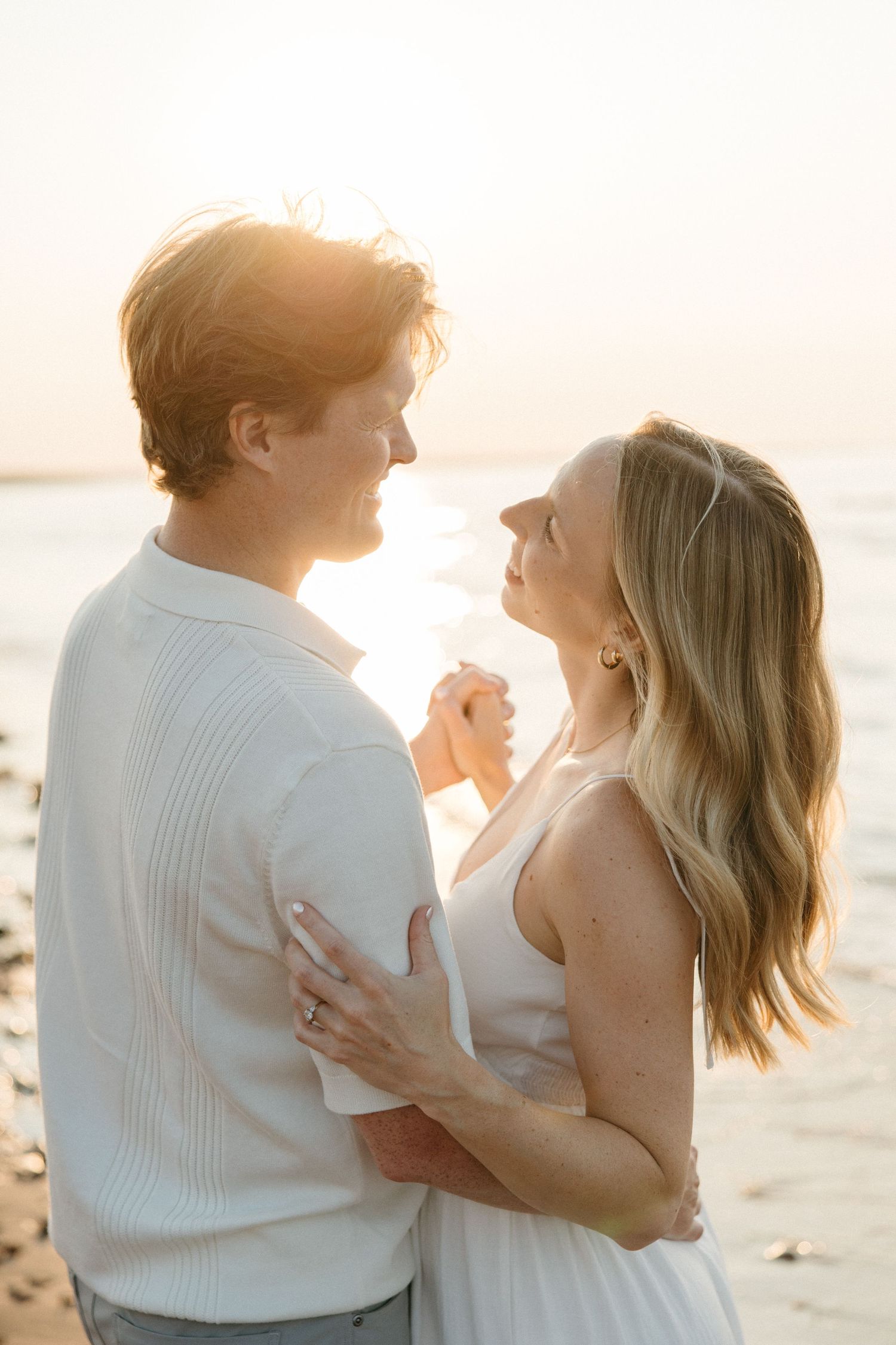 Smiling couple embracing and holding hands on a sunlit beach during golden hour by the sea.