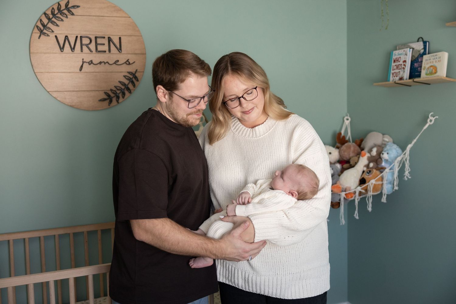 Parents pose with their newborn in a nursery with sage green walls and wooden decor.
