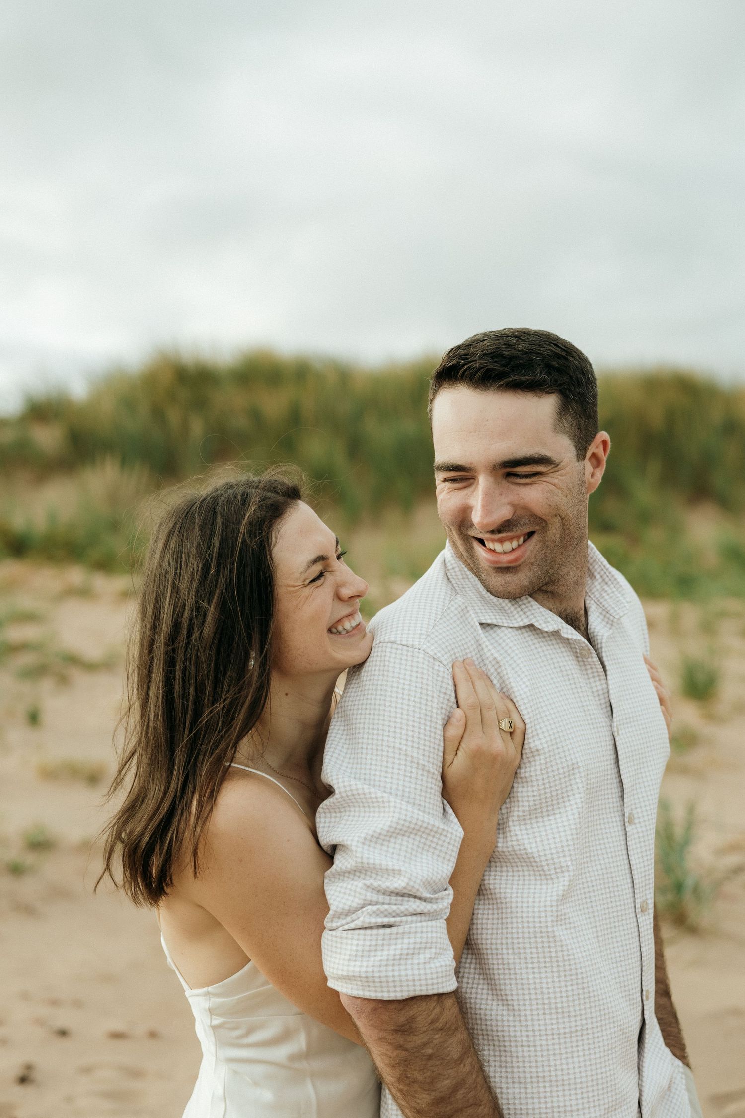 A couple shares a candid laughing moment together on a beach with dunes in the background.