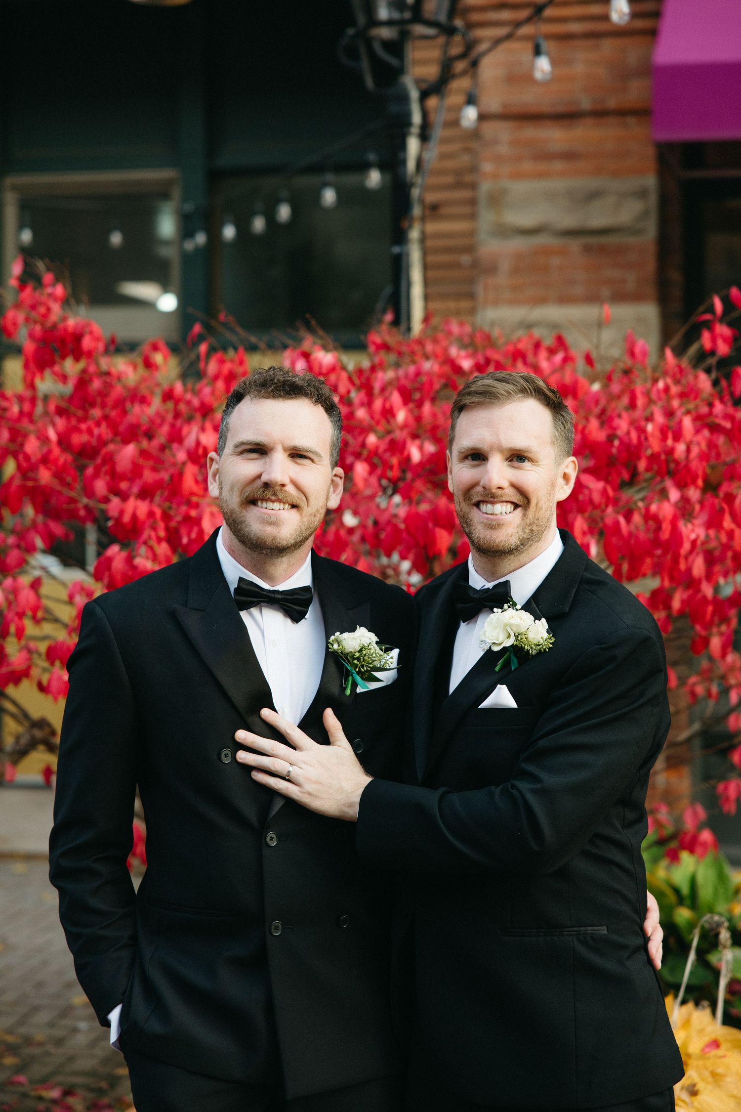 Groomsmen in black tuxedos share a candid moment against vibrant red flowers.