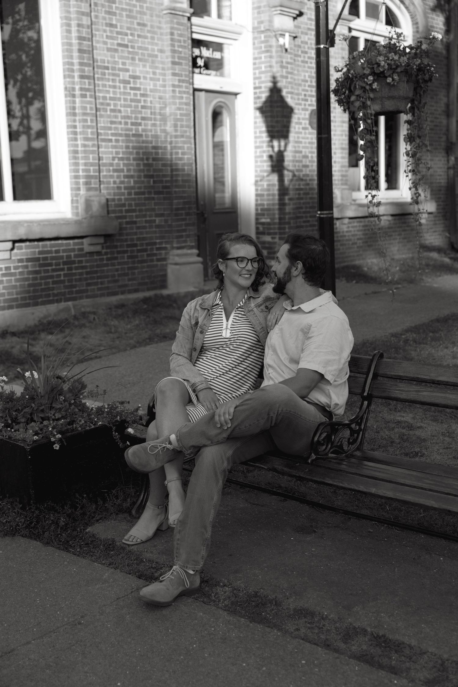 A couple sits together on a bench outside a historic brick building in black and white.