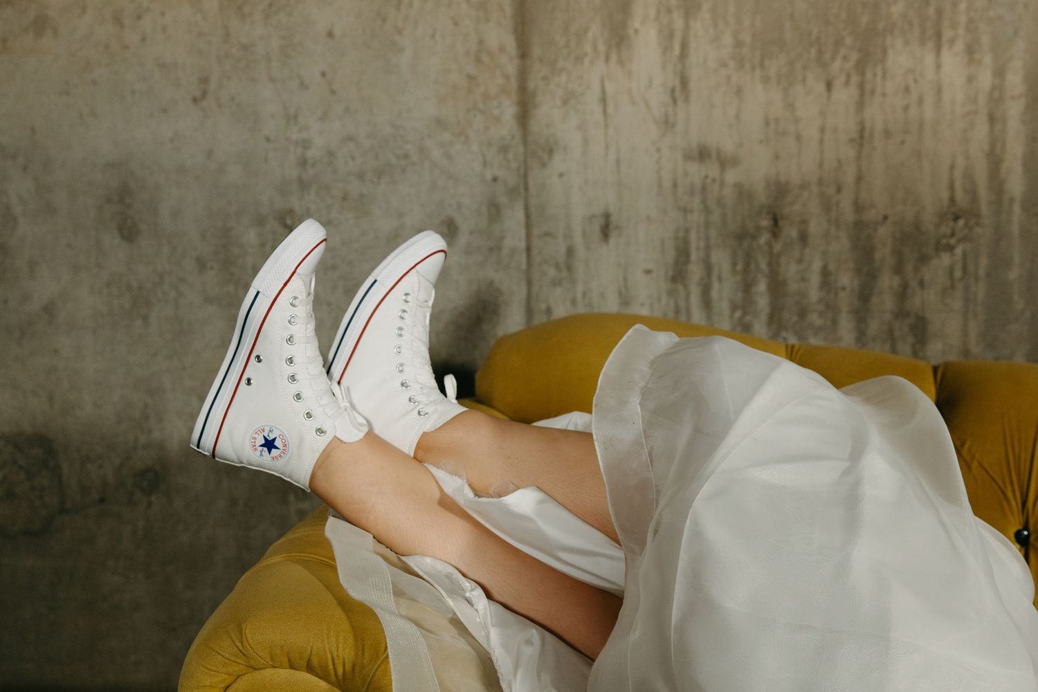 Close-up of white sneakers worn with wedding dress while lounging on yellow couch.