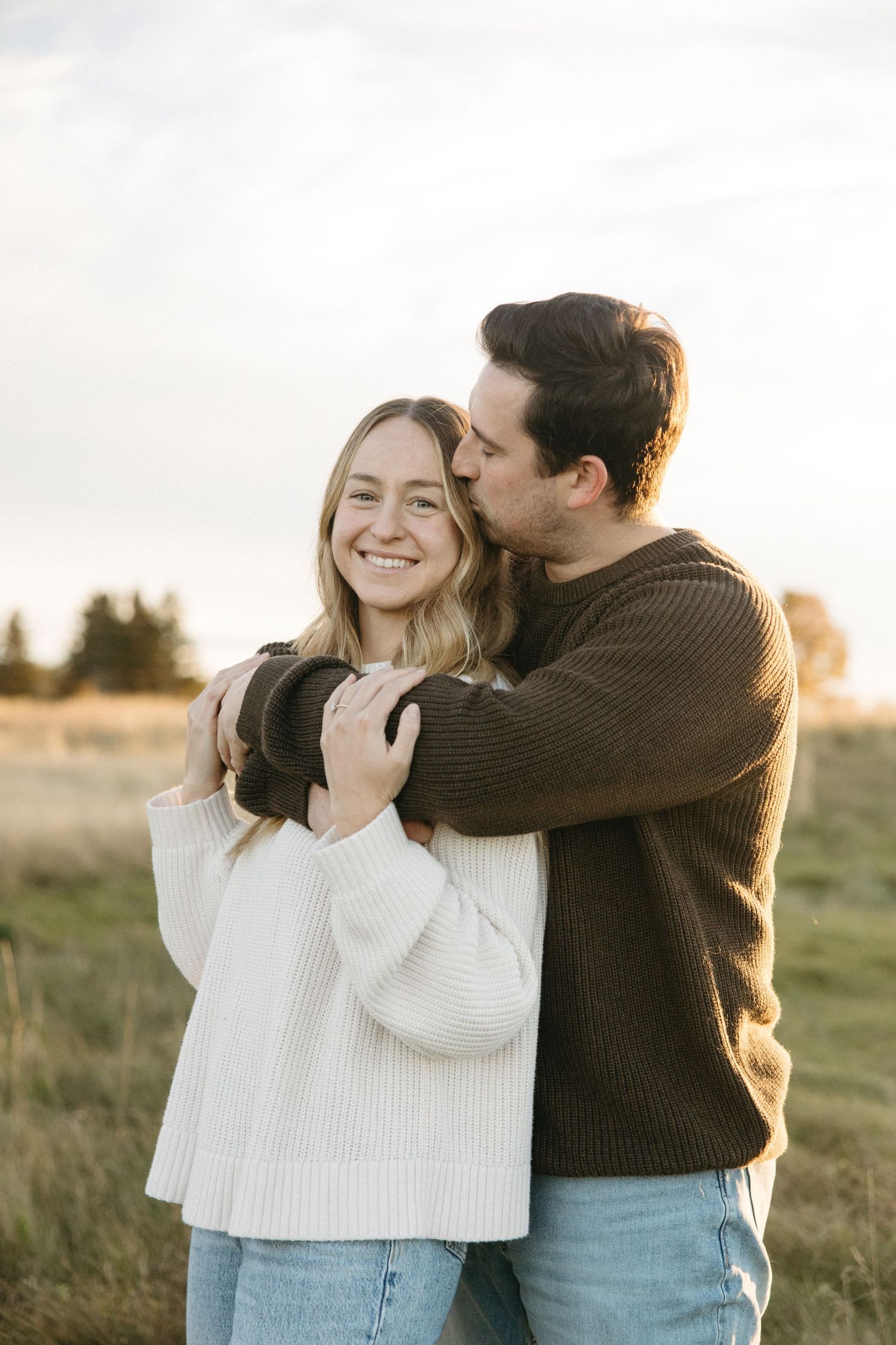 Smiling woman in a cream knit sweater embraced from behind by a man in a brown sweater kissing her cheek in a golden grassy field.