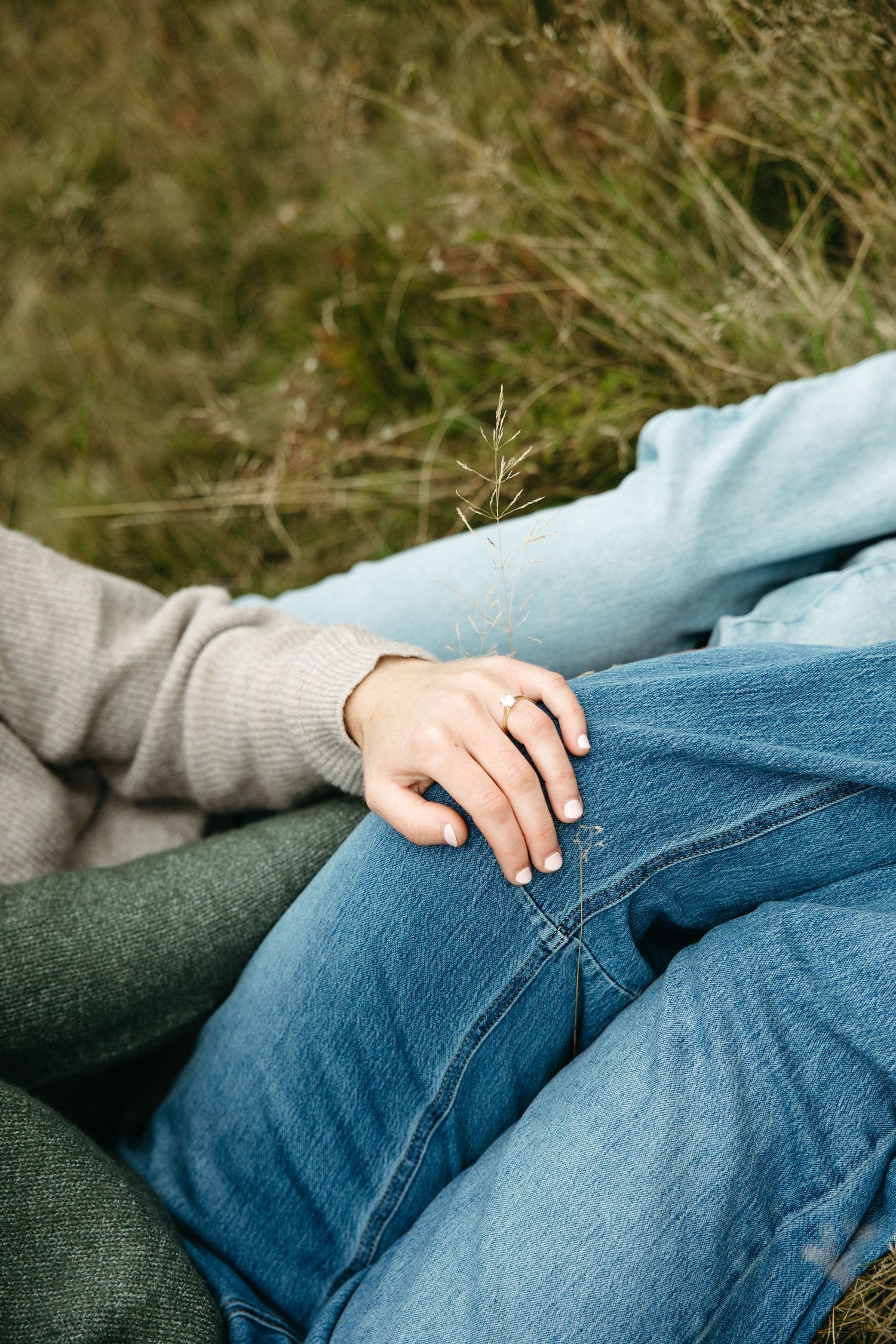 Close-up of a hand with a ring resting on denim-clad knees as two people sit together on the grass in a field.
