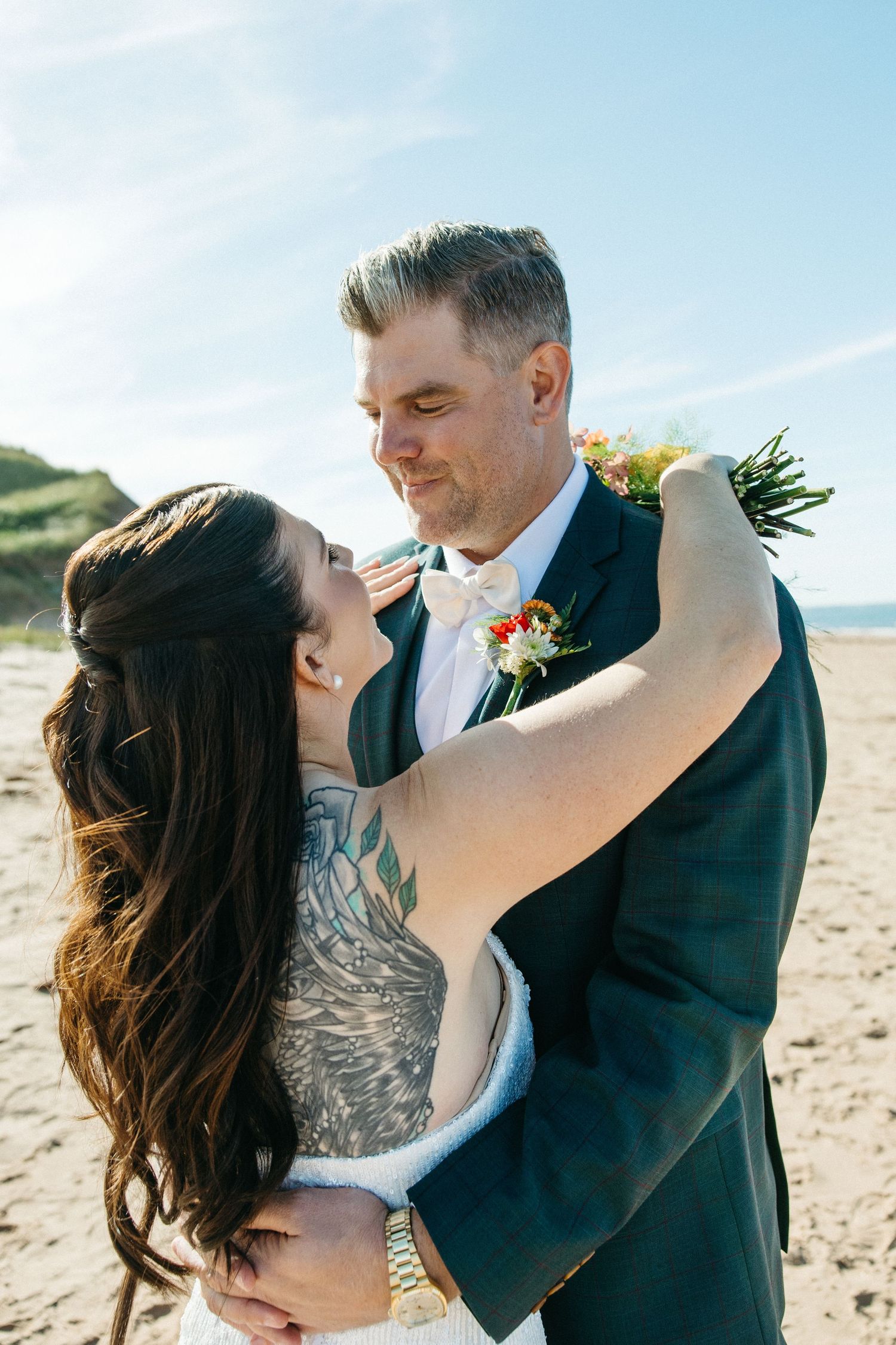 Groom in a navy suit and bow tie hugs his tattooed bride on a sunny beach with blue sky and dunes.
