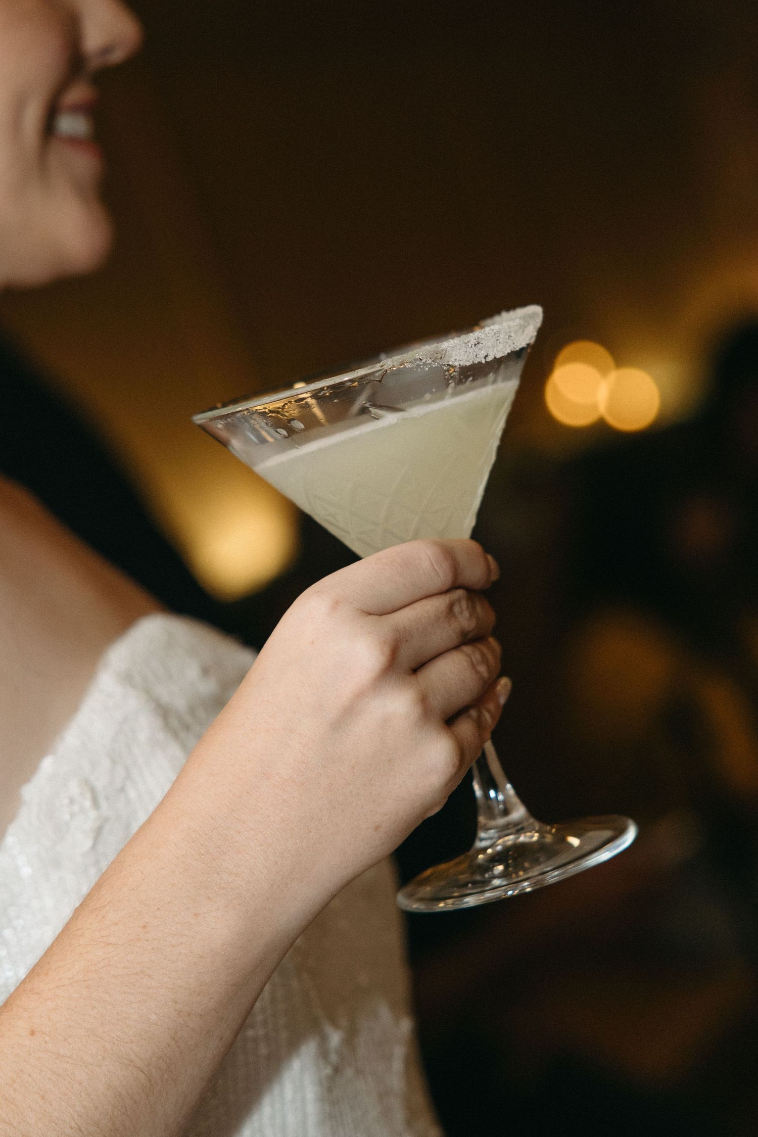 Woman in a white lace dress holds a sugar-rimmed martini glass filled with a pale green cocktail at a warmly lit evening event.