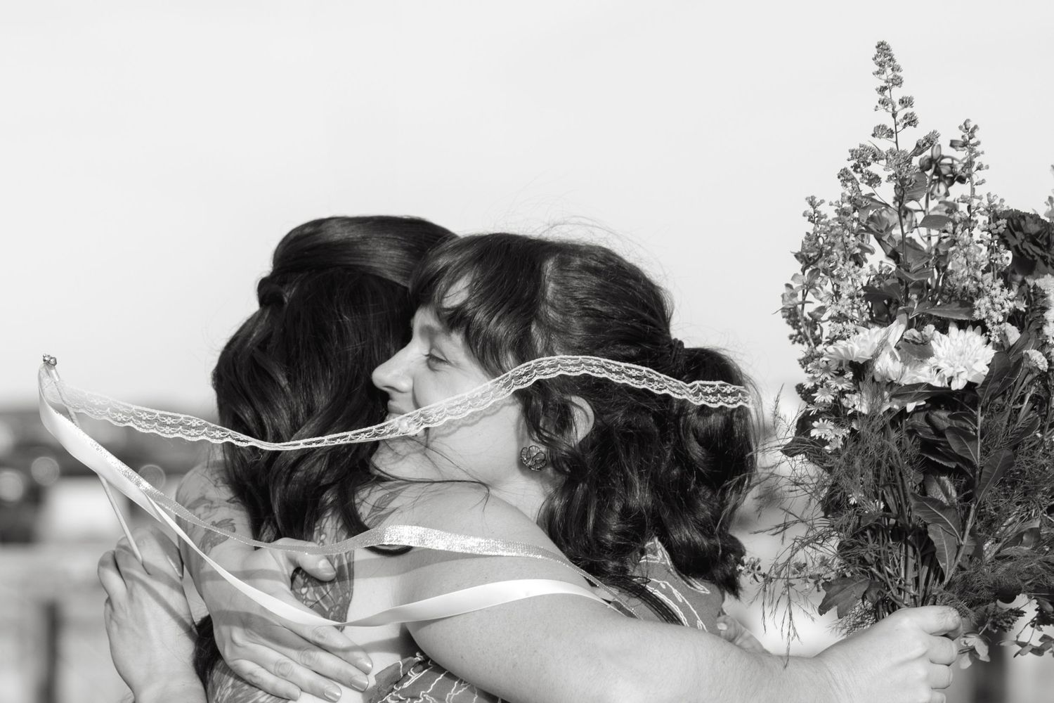 Two women with dark hair share a warm embrace while one holds a colorful bouquet of flowers in this black and white photograph.