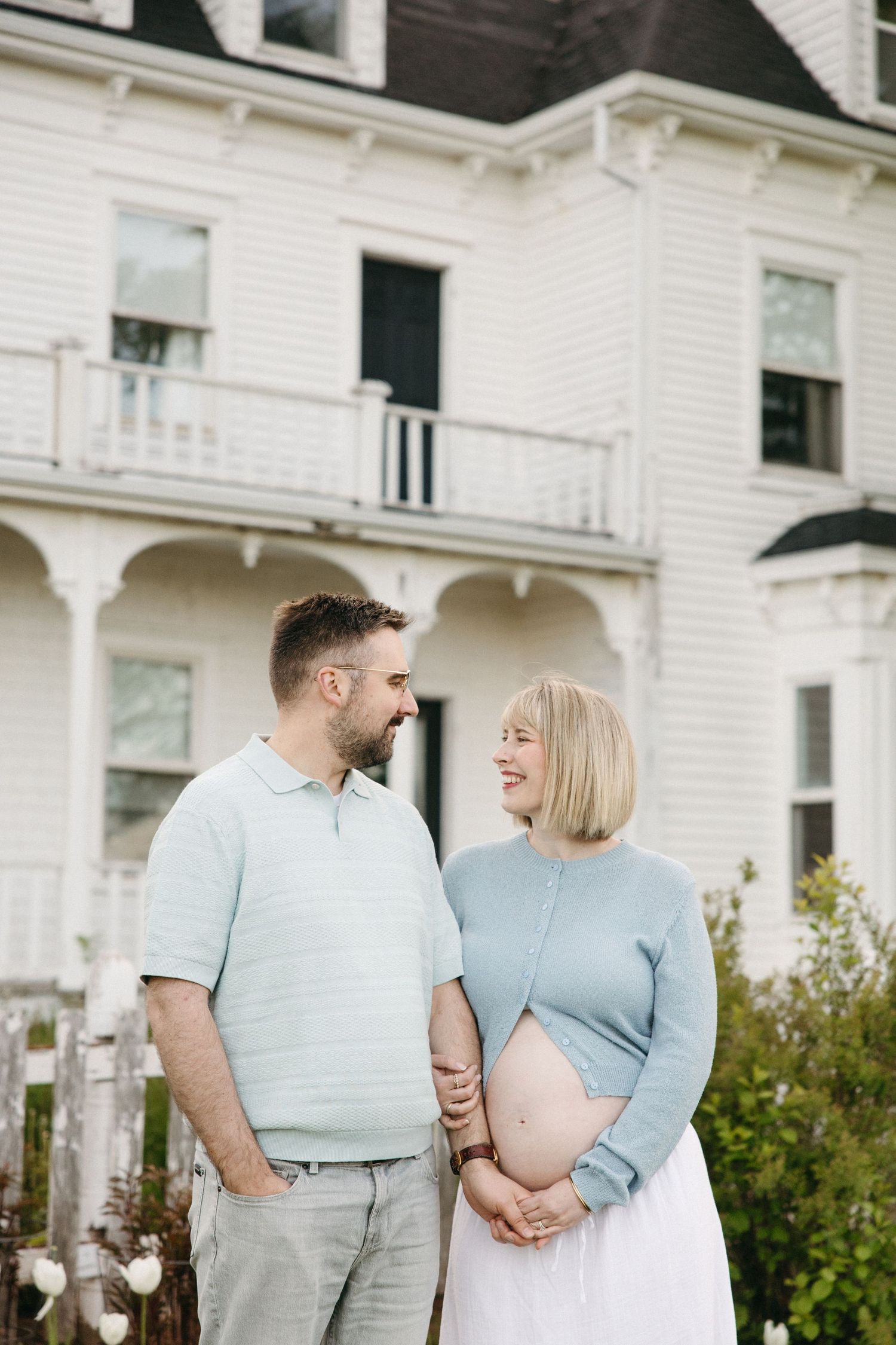 Pregnant couple smiling at each other in front of a white Victorian house, wearing matching blue outfits.