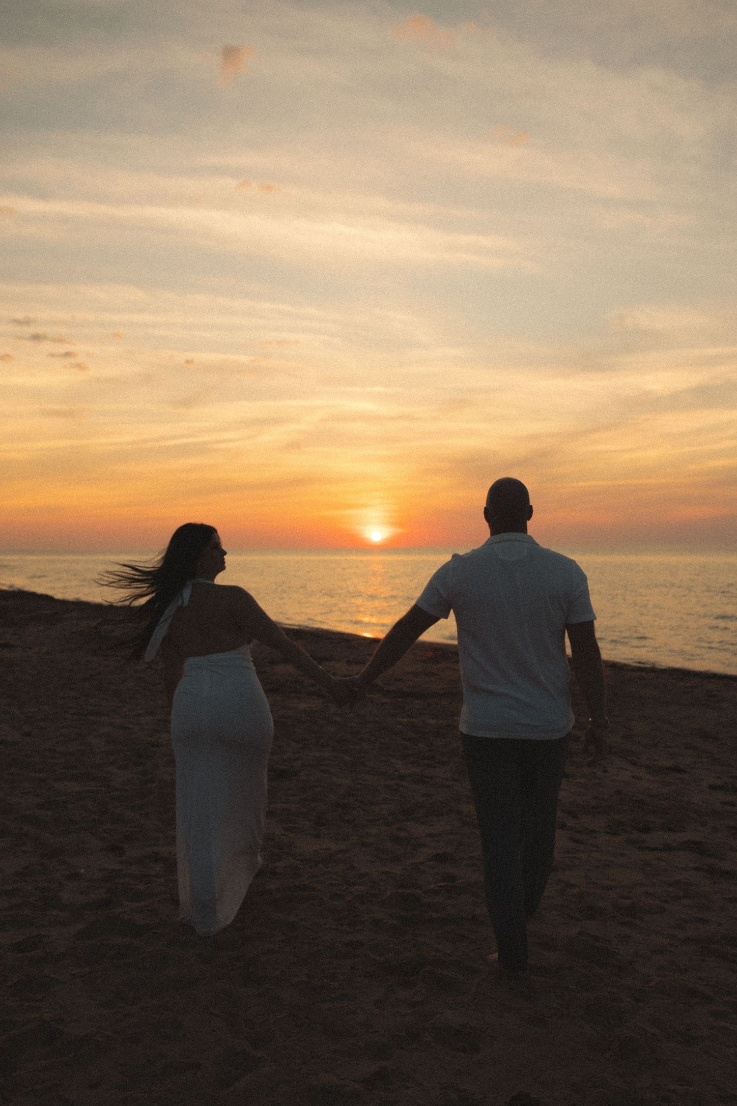 Couple holding hands walking toward stunning ocean sunset on a sandy beach.