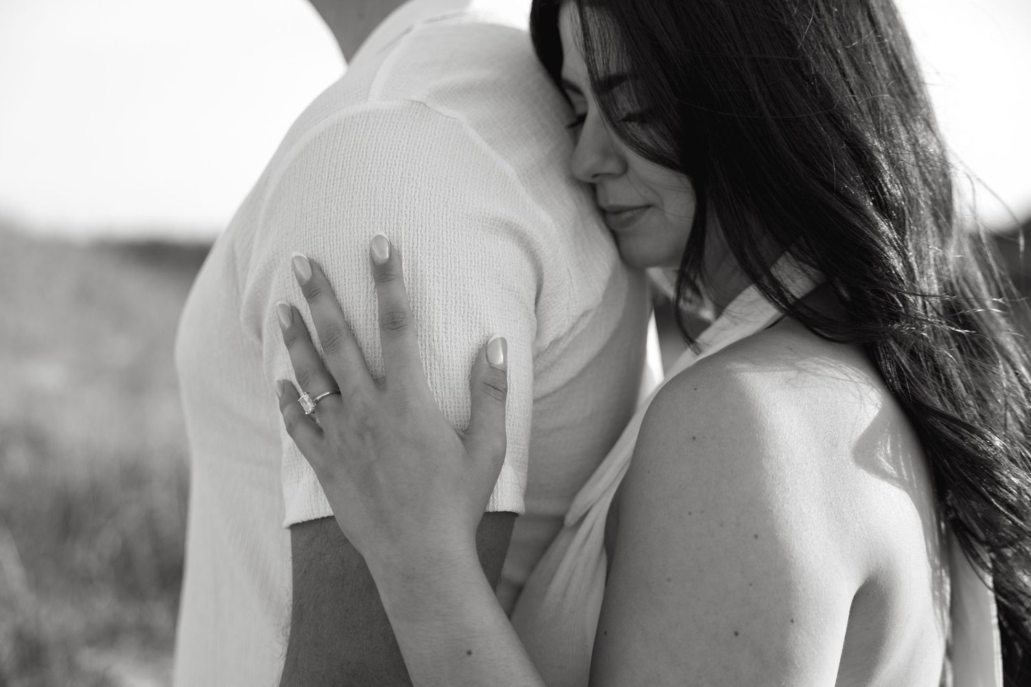 Black and white photo of a couple embracing, woman with engagement ring resting her head on mans shoulder.