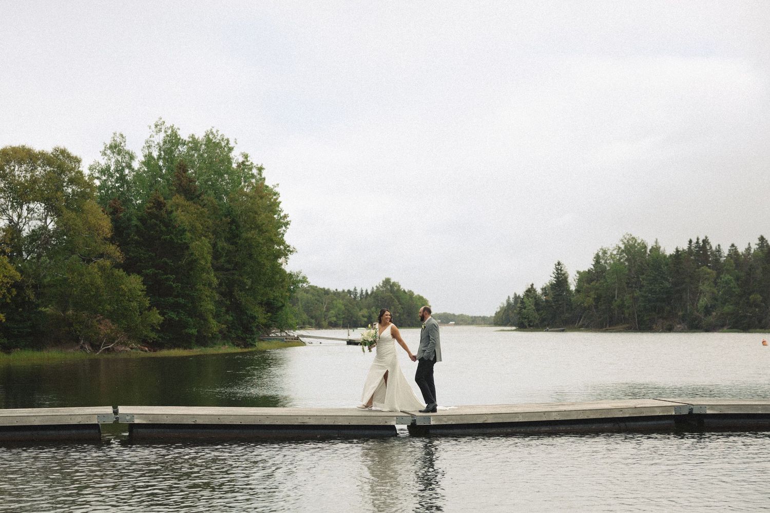 A couple poses for photos on a scenic dock overlooking a lake surrounded by trees.