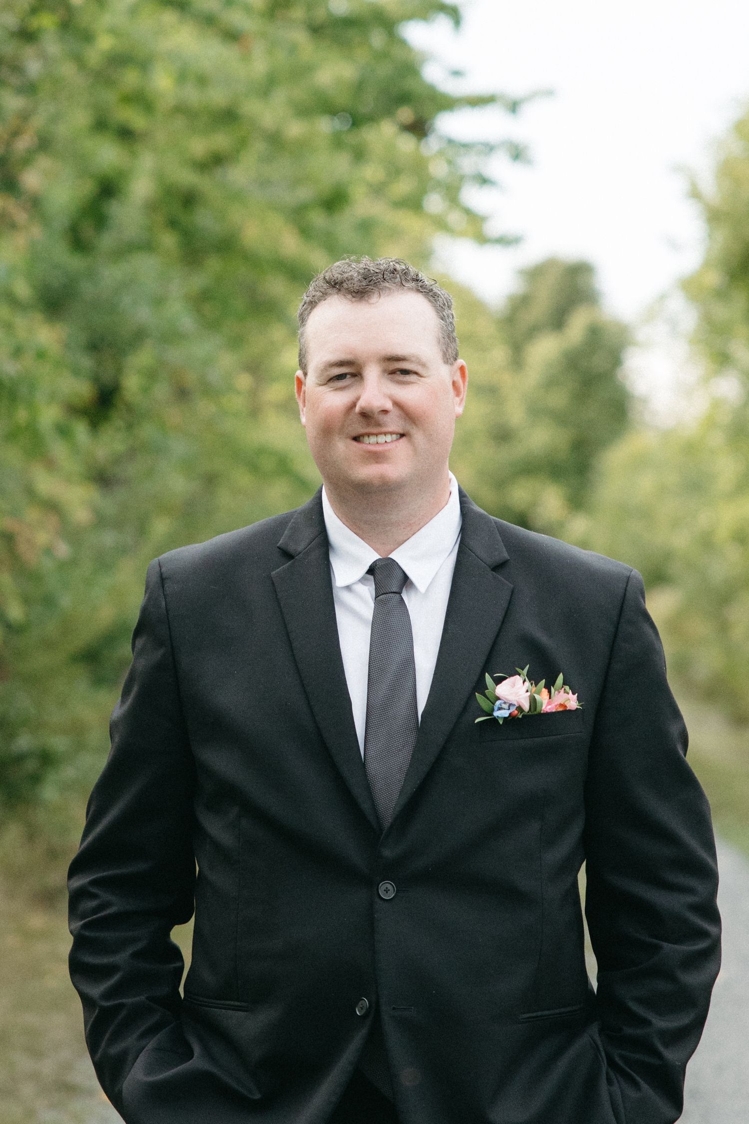 Groom in black suit and tie smiling outdoors with trees in background.