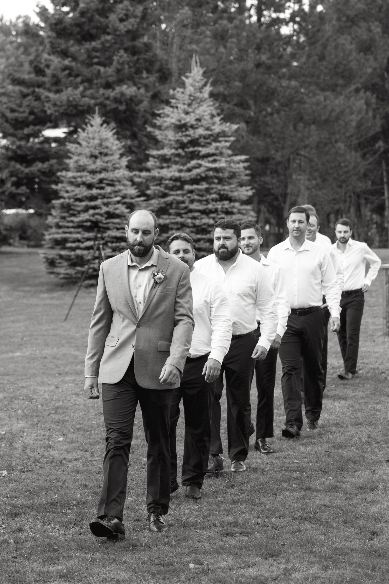 Groomsmen walking in a line across a grassy lawn during wedding photos.