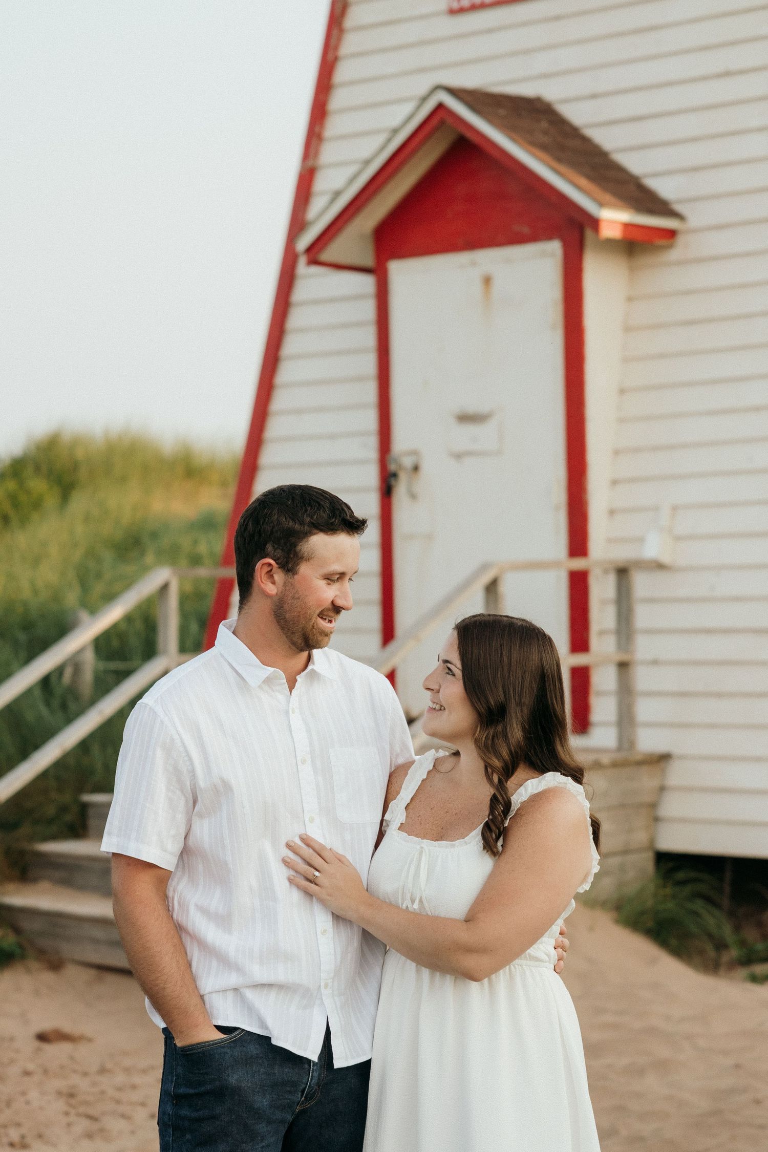 Happy couple laughing together near a red and white lighthouse on a beach.
