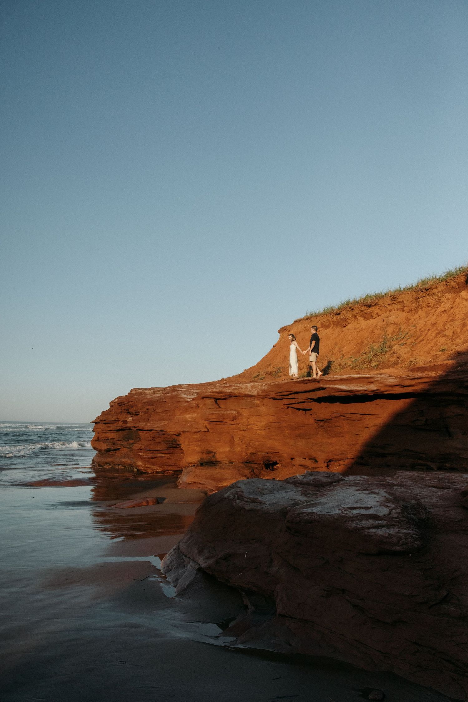 A couple stands on red coastal cliffs overlooking the ocean at sunset.
