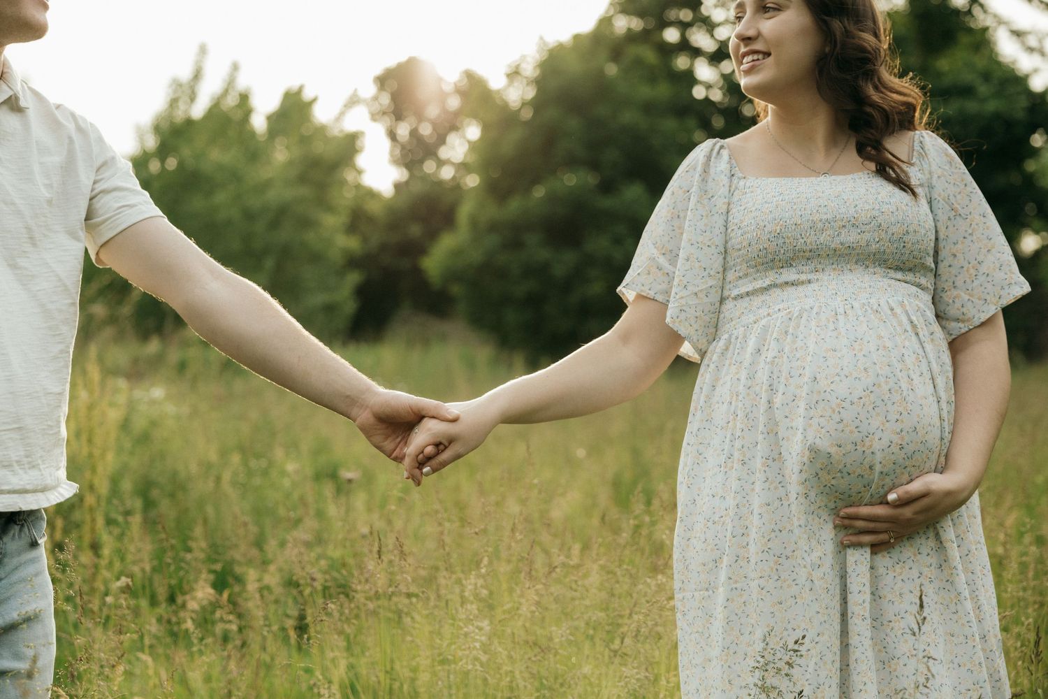 A couple in light-colored clothing embraces while standing in a grassy summer field at golden hour.