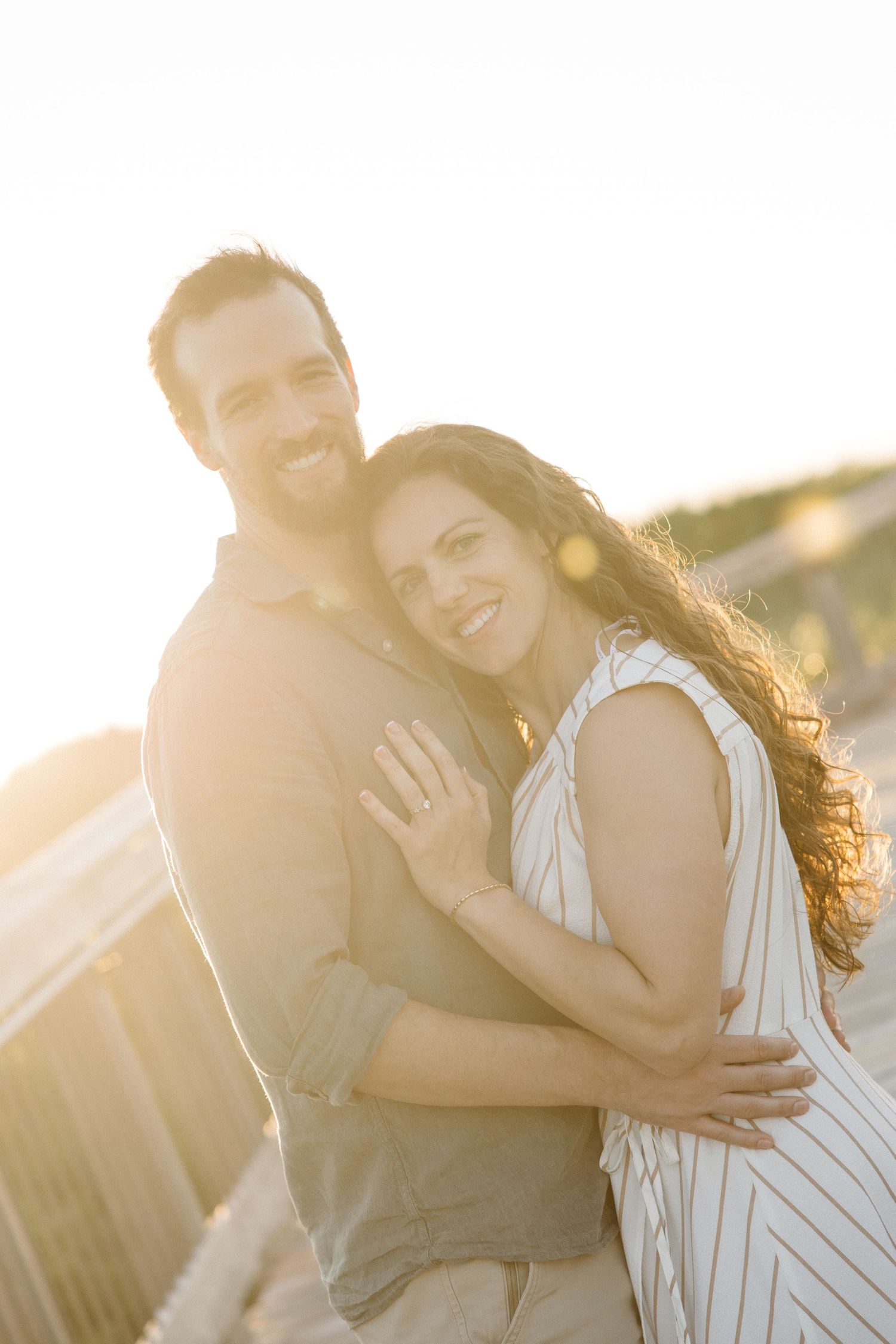 Couple embracing and smiling in warm golden sunset backlight, woman showing engagement ring.