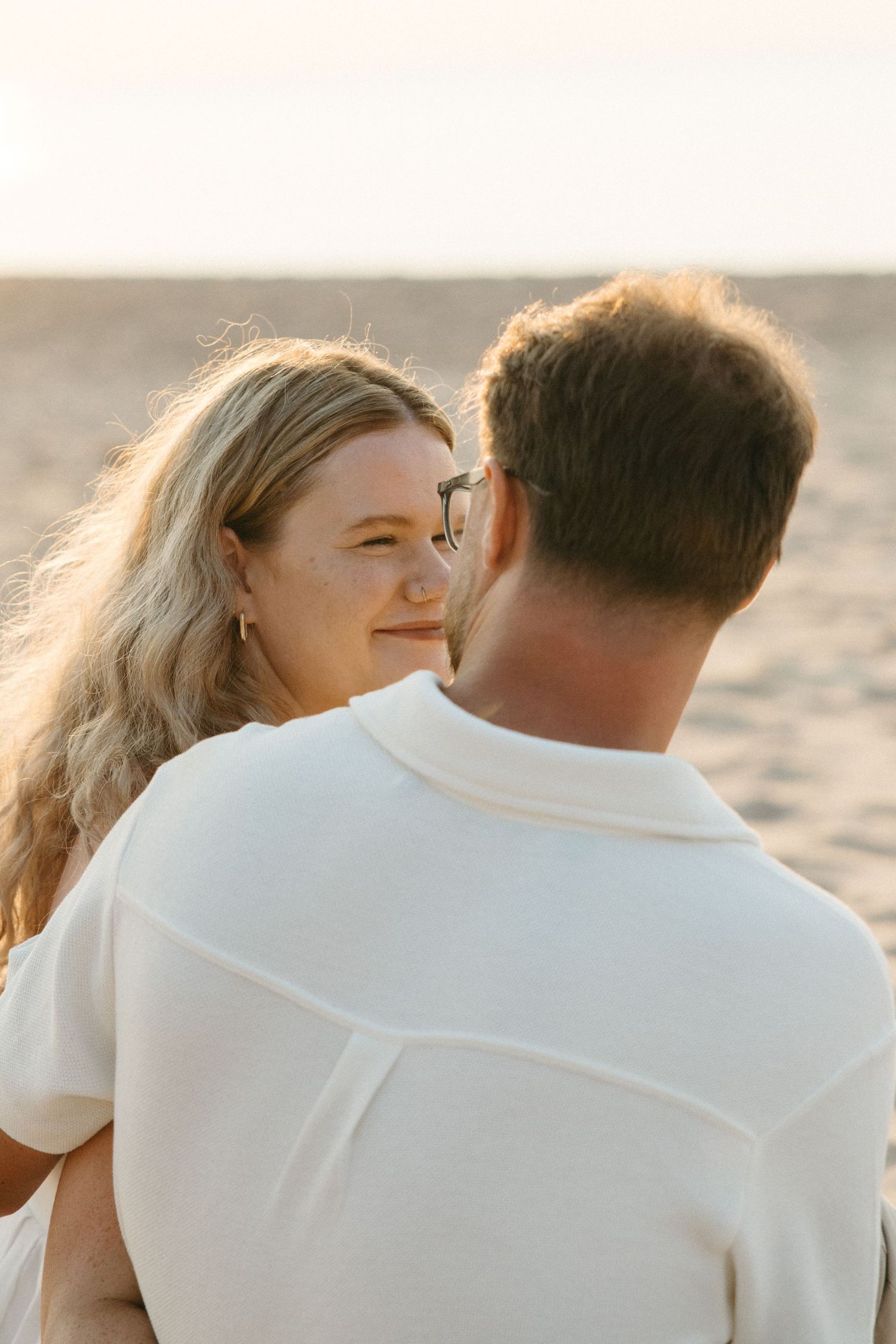 Couple embracing on a sandy beach at golden hour, woman with blonde hair smiling warmly at man in white polo shirt.