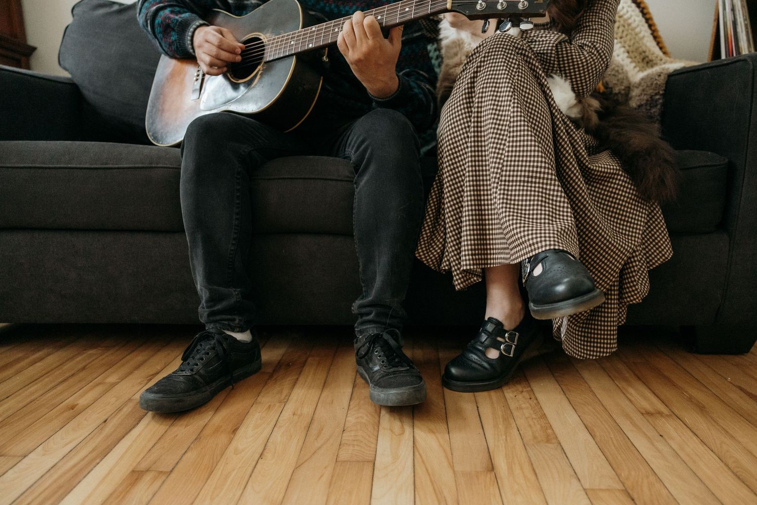 Two people sitting on a couch, one playing acoustic guitar, wearing black shoes on hardwood floor.