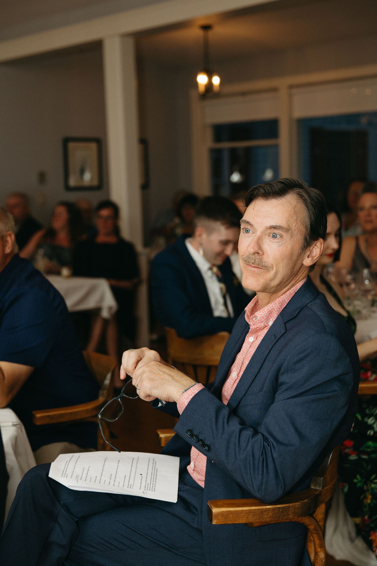 Man in navy suit holding glasses and program seated at wedding ceremony.