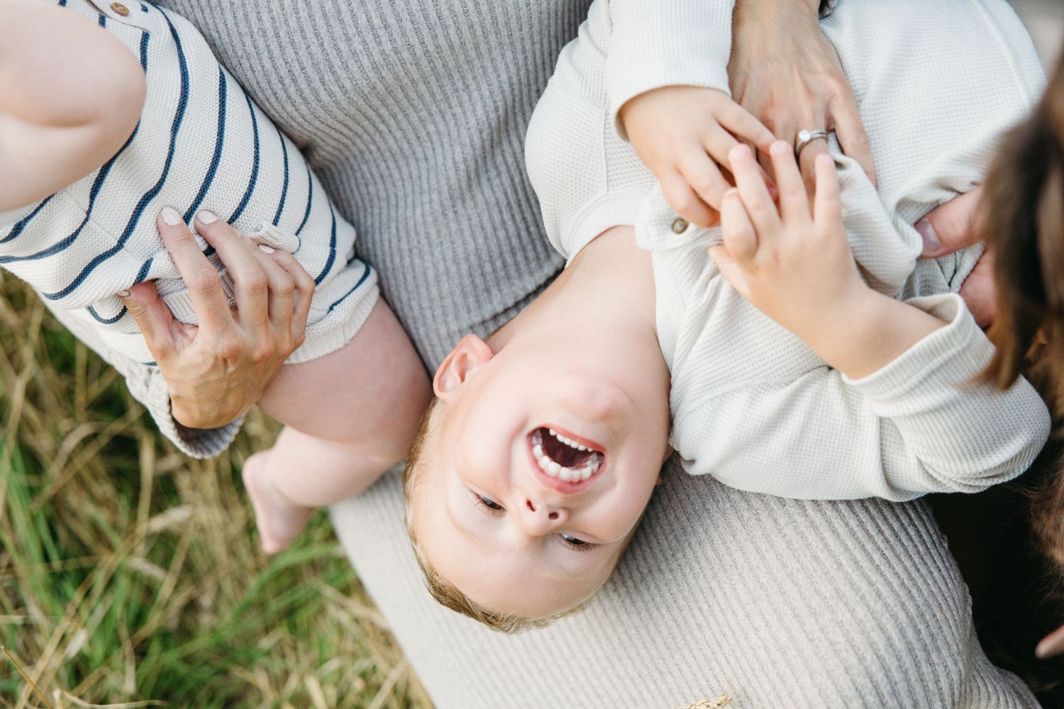 Happy baby laughing upside down while being held by parents outdoors in a grassy field.