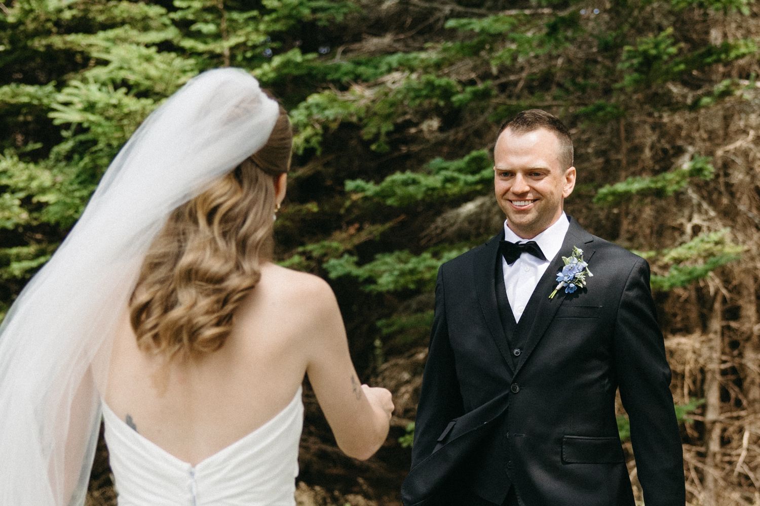 Groom in black tuxedo smiling at bride during emotional first look moment in a forest setting.
