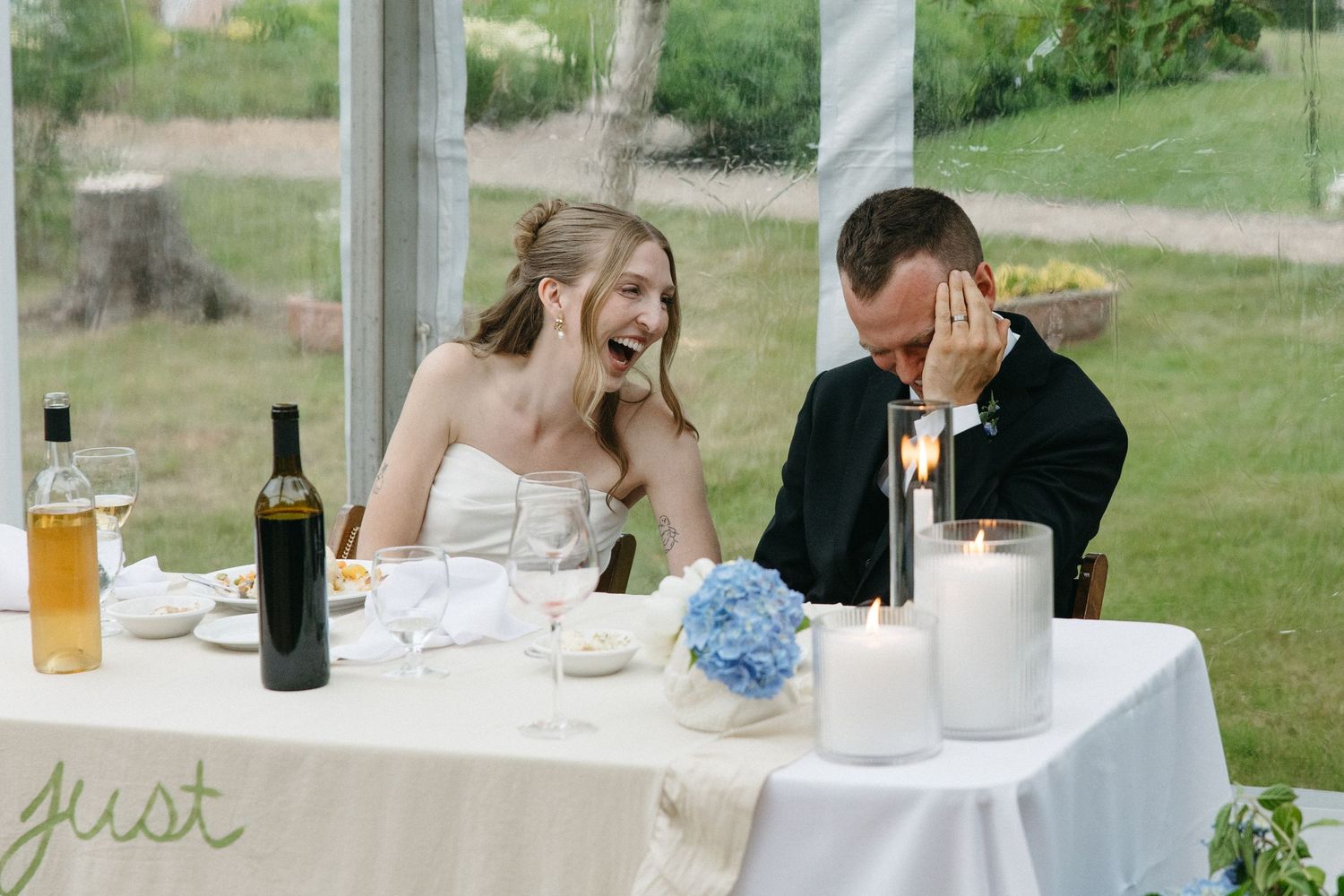 Bride laughing joyfully with groom at sweetheart table during wedding reception speeches.