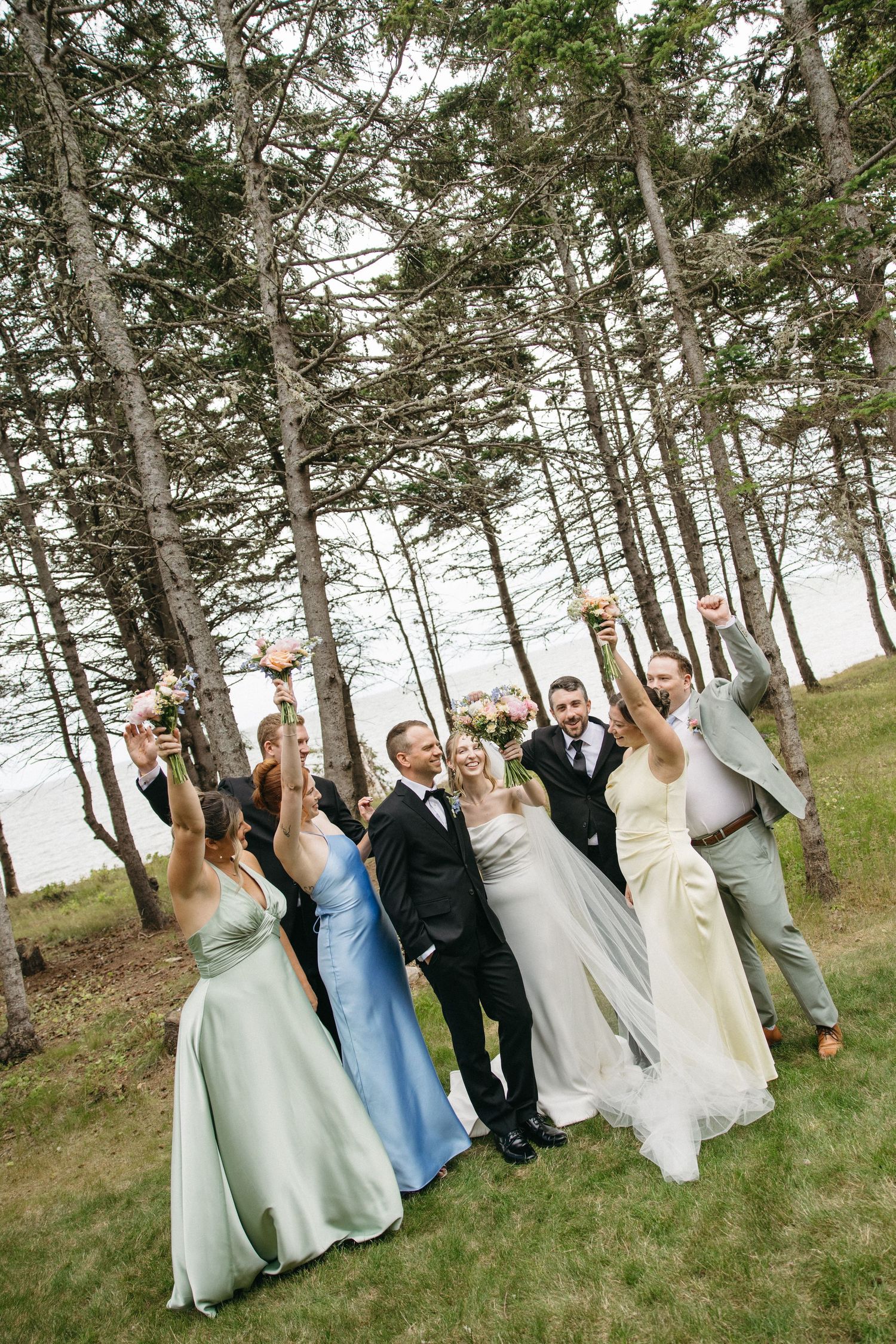 Joyful wedding party celebrating outdoors among tall pine trees near the ocean, bridesmaids in pastel gowns raising bouquets.