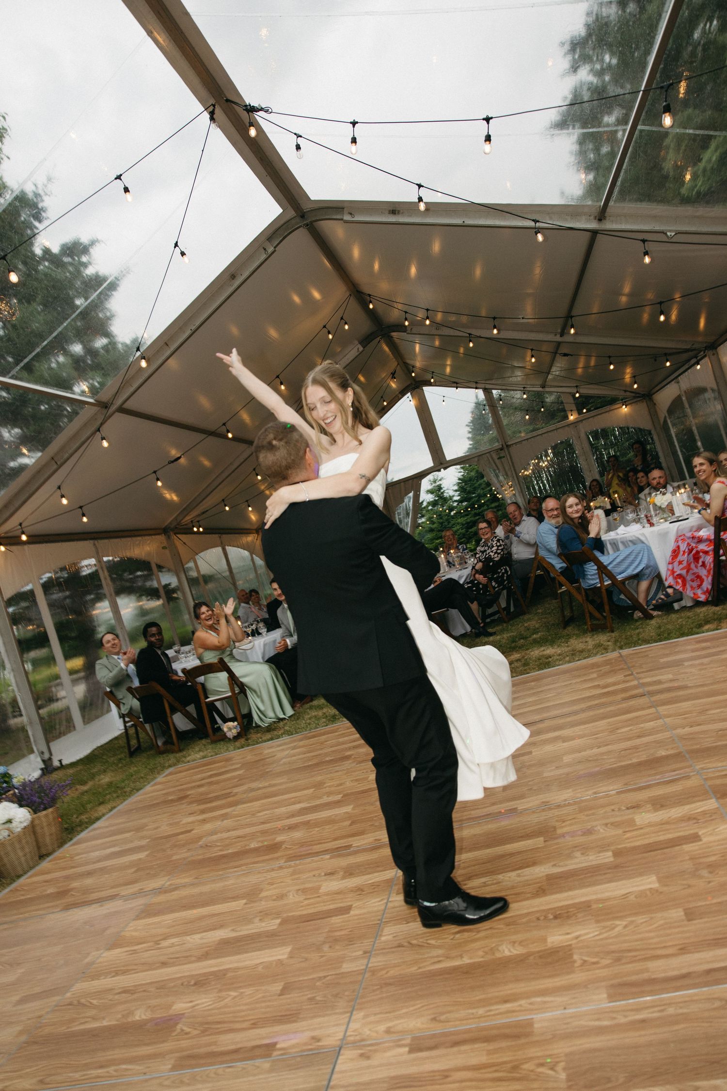 Groom lifting bride during first dance under clear tent with string lights.