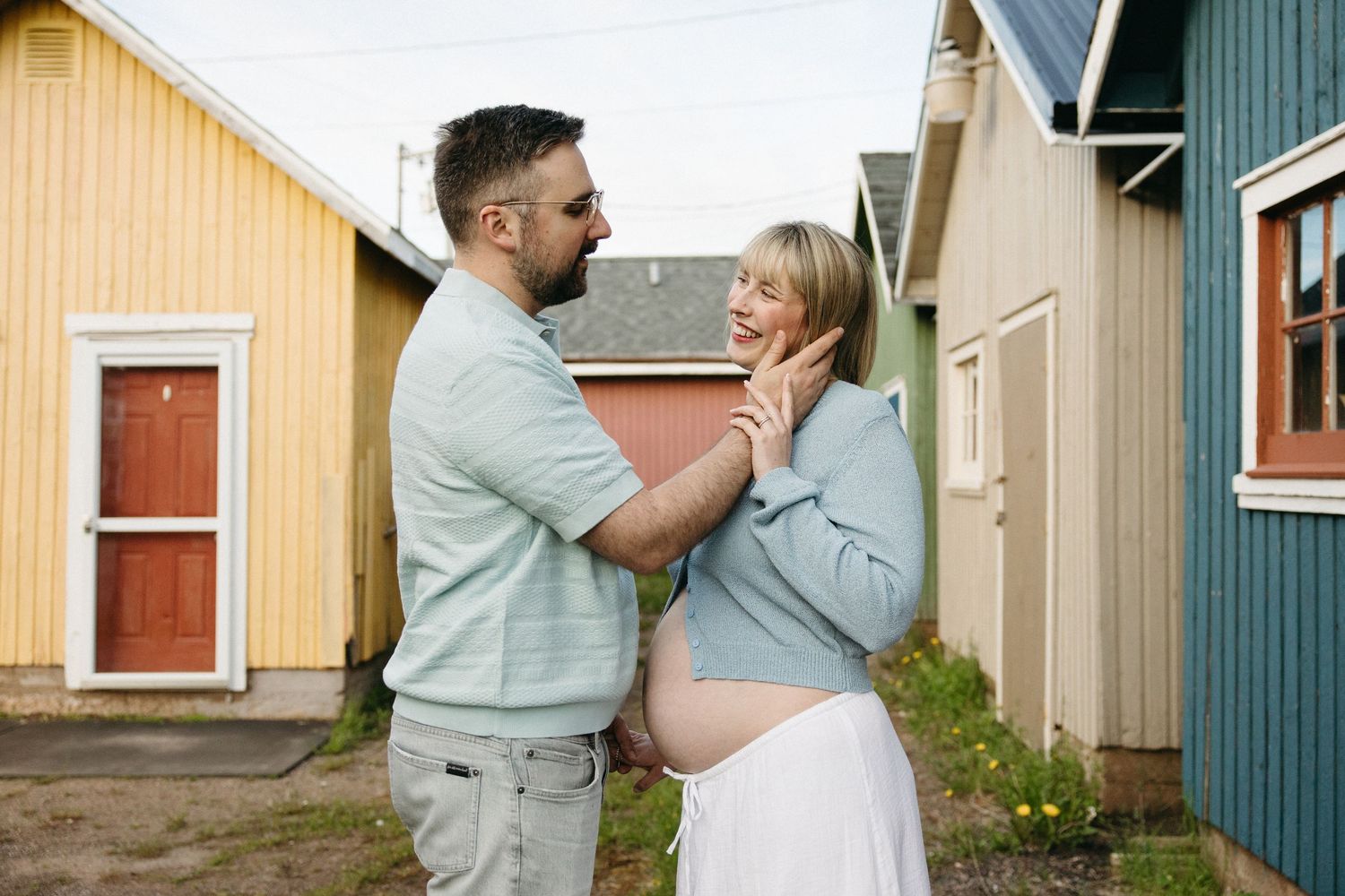 Man tenderly cupping pregnant woman's face between vibrant colored wooden buildings during maternity shoot.