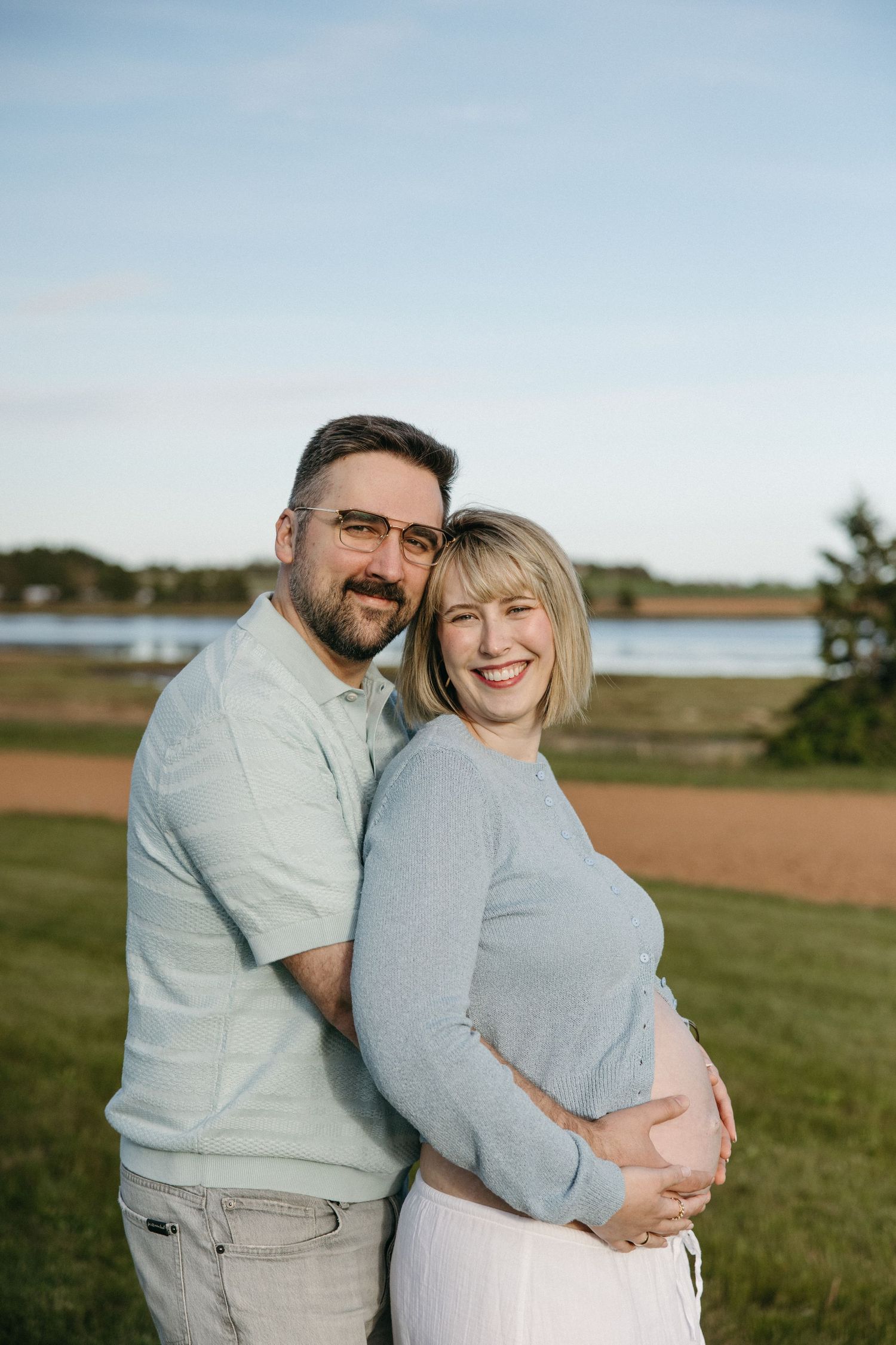 Smiling couple posing outdoors near a lake, woman cradling pregnant belly, wearing light blue outfits on a sunny day.
