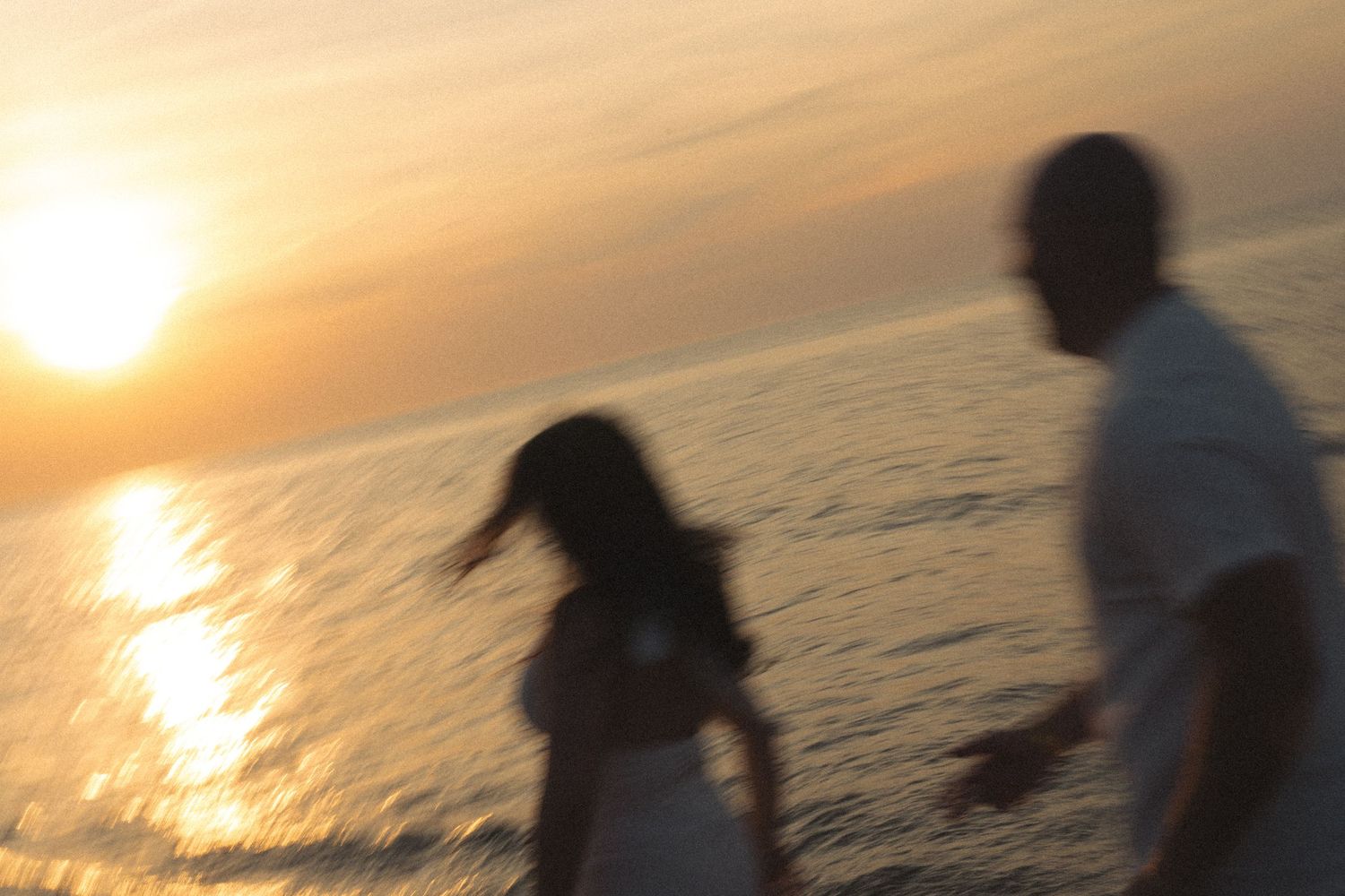 Blurry couple holding hands on a beach at sunset with golden light reflecting on the ocean water.