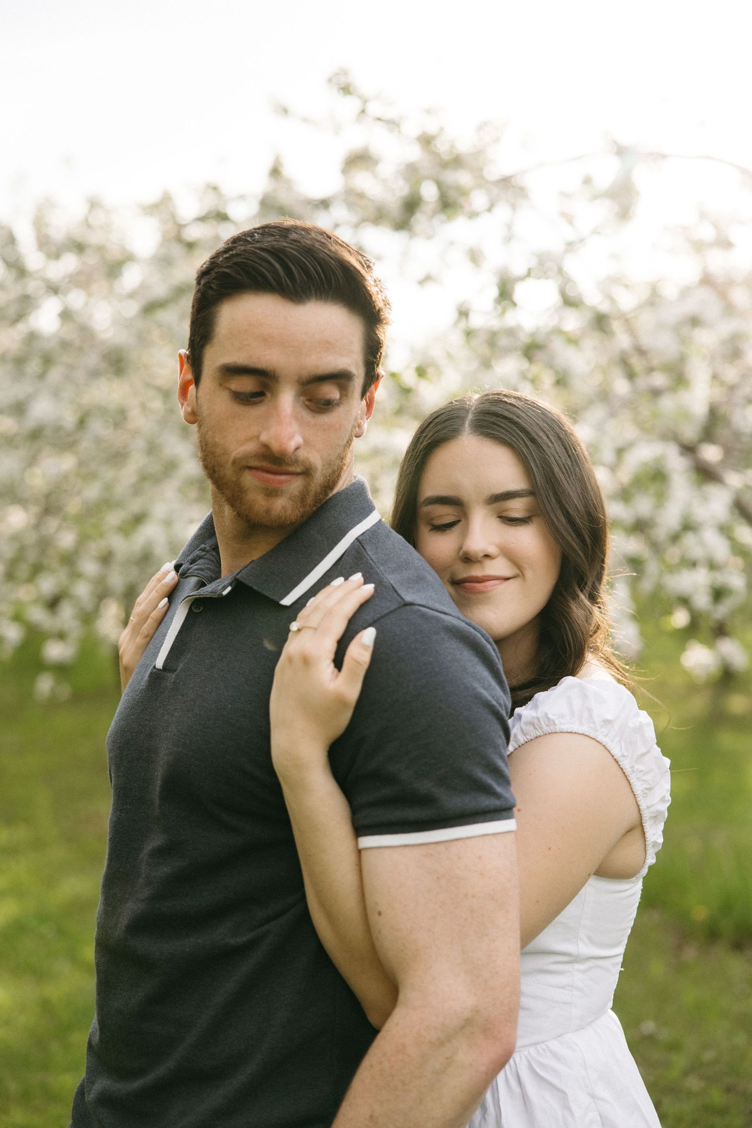 Woman in white dress embracing man in navy polo shirt among white flowering trees in spring.