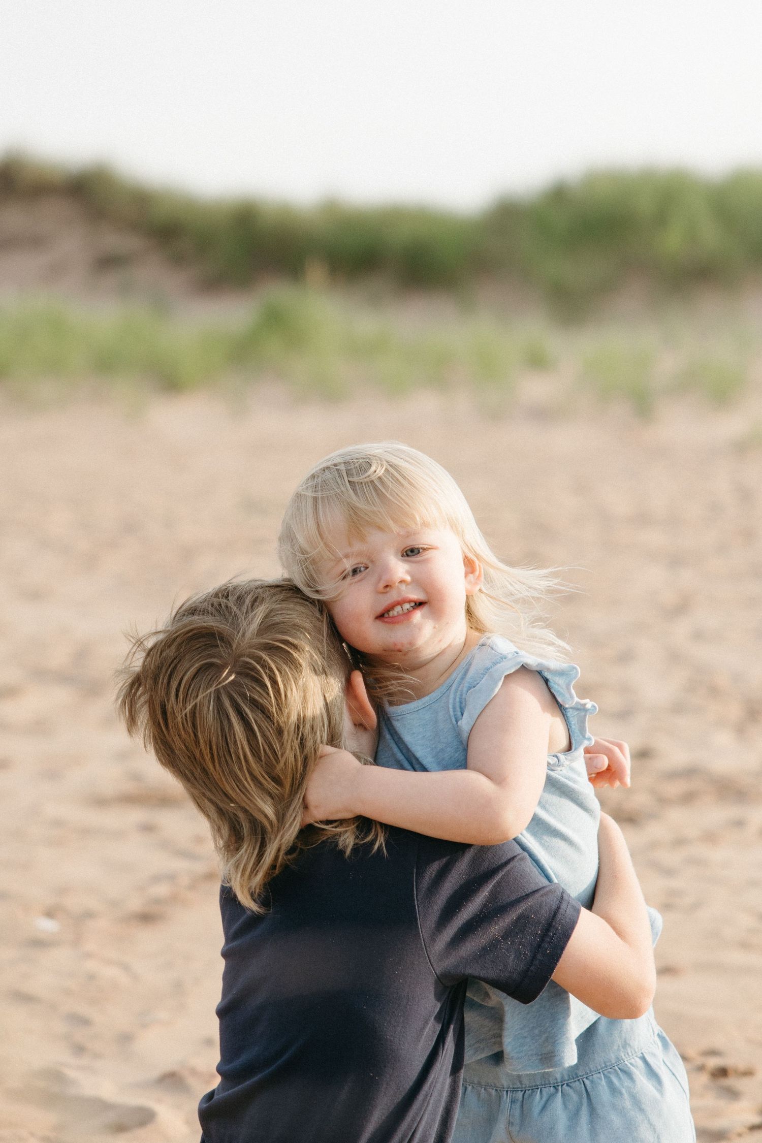 Mother hugging smiling toddler girl in blue dress on a sunny beach.
