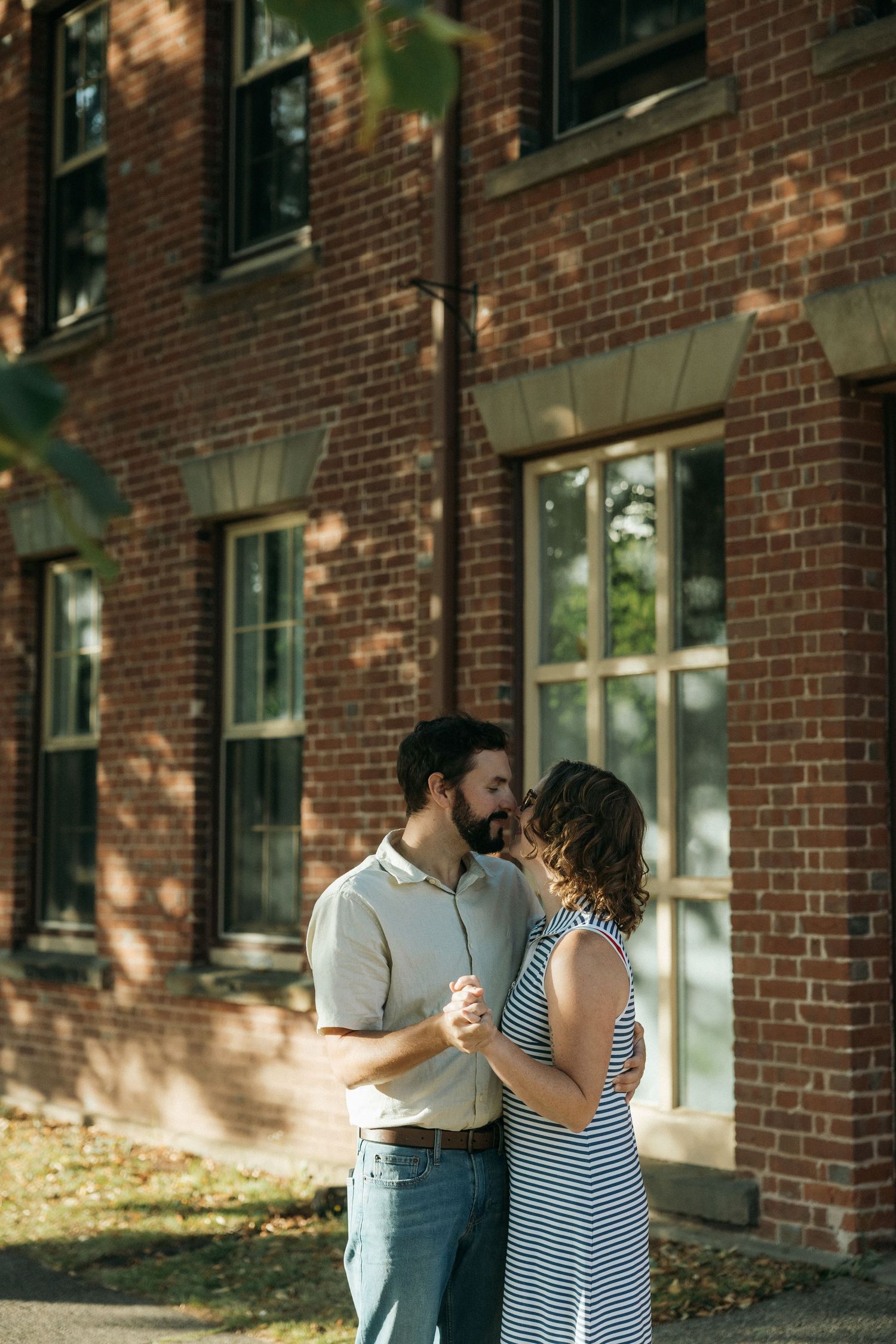 A couple shares an intimate moment outside a historic brick building in warm sunlight.