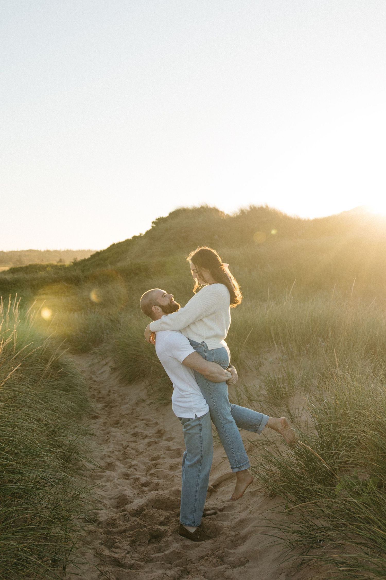 A couple dances and spins playfully in a grassy field during sunset.