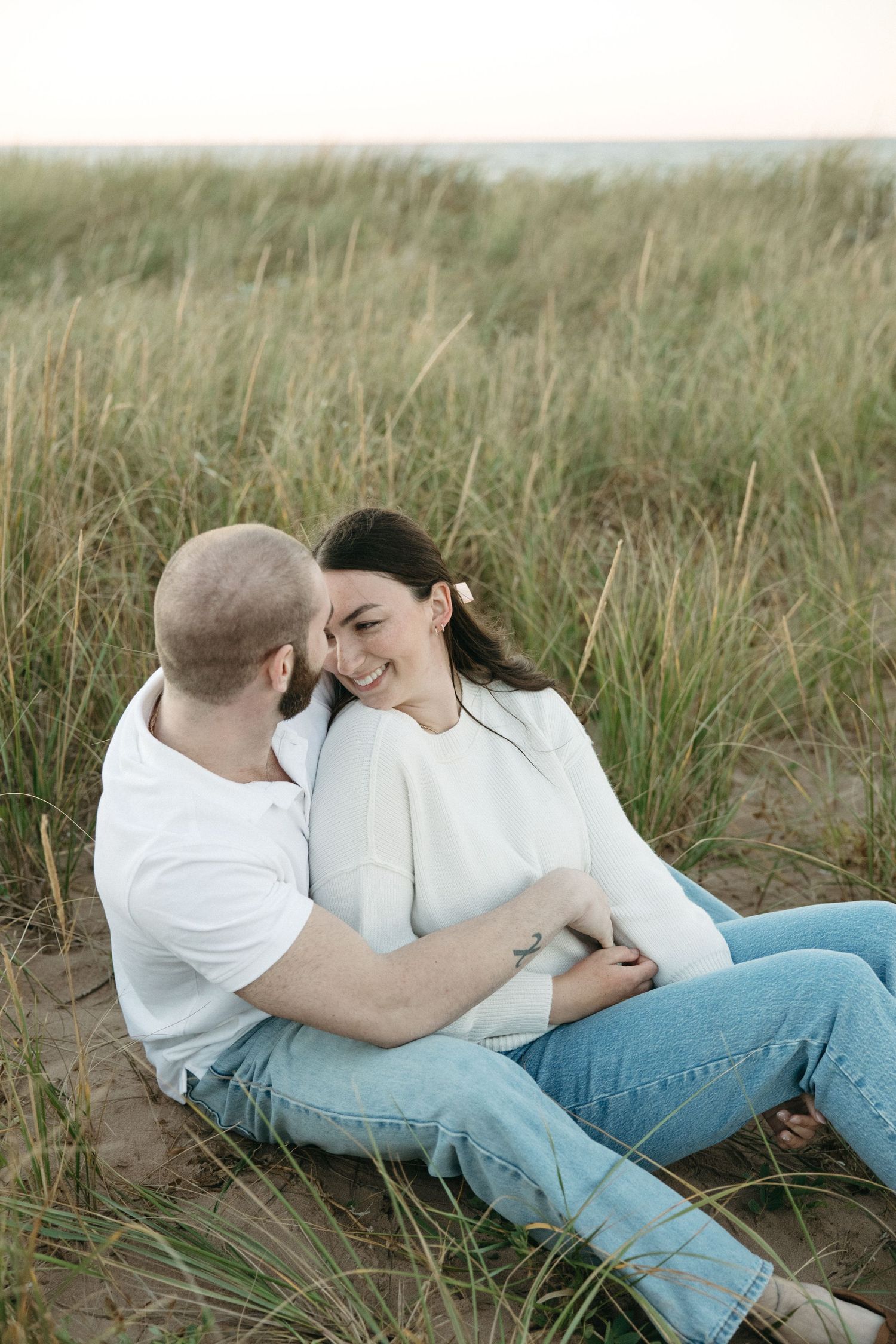 A romantic couple lying in tall grass wearing white shirts and blue jeans in a natural outdoor setting.