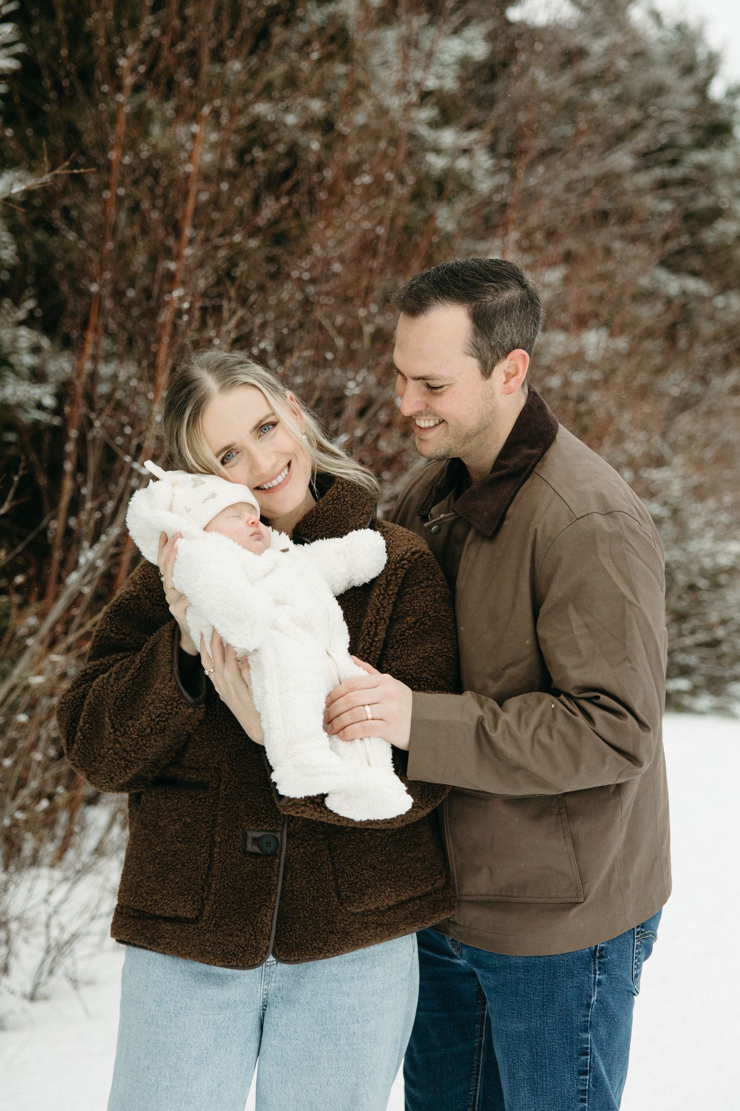 Family sharing intimate winter moment with baby in white snowsuit.