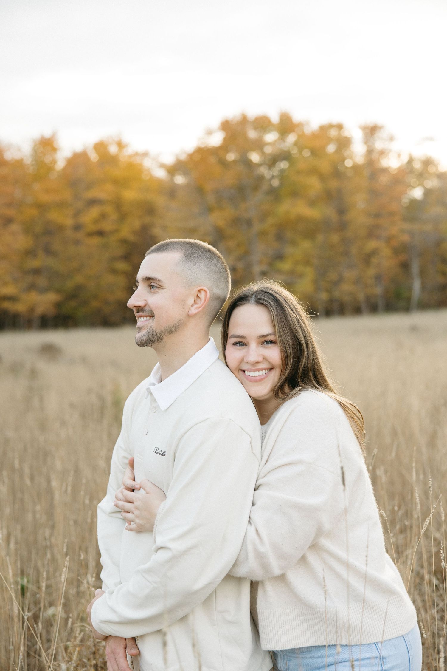 Couple in matching cream sweaters embracing in autumn field with golden trees in background.