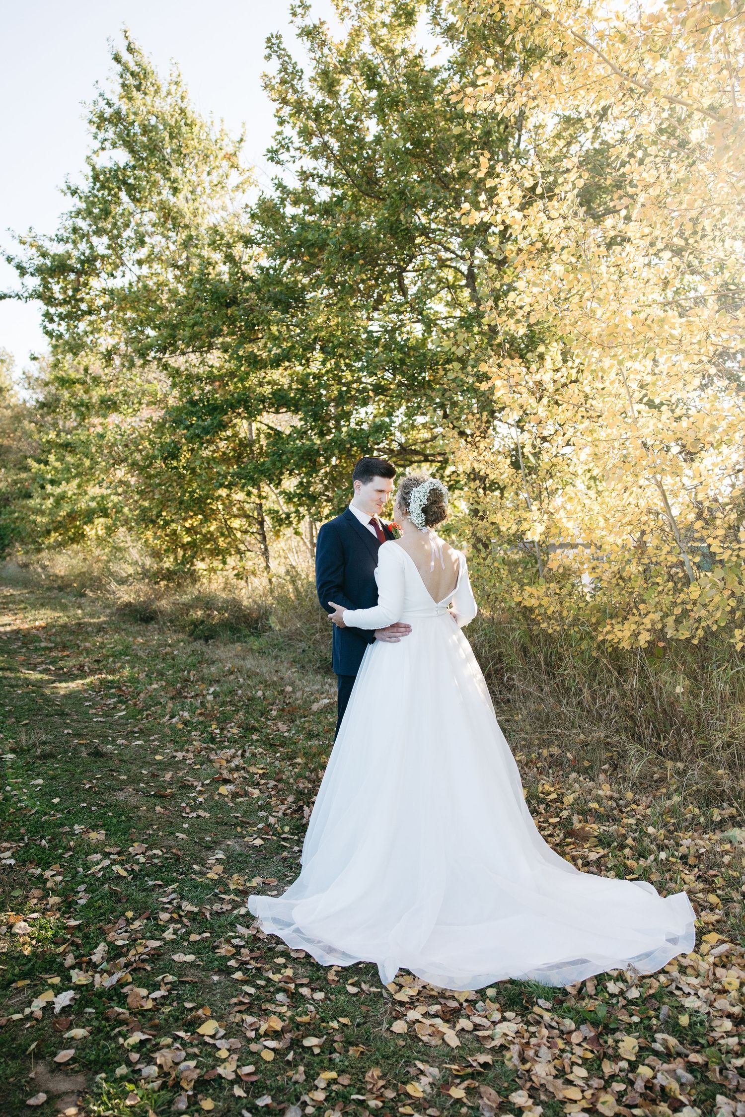 A couple in wedding attire embrace among autumn trees and fallen leaves.