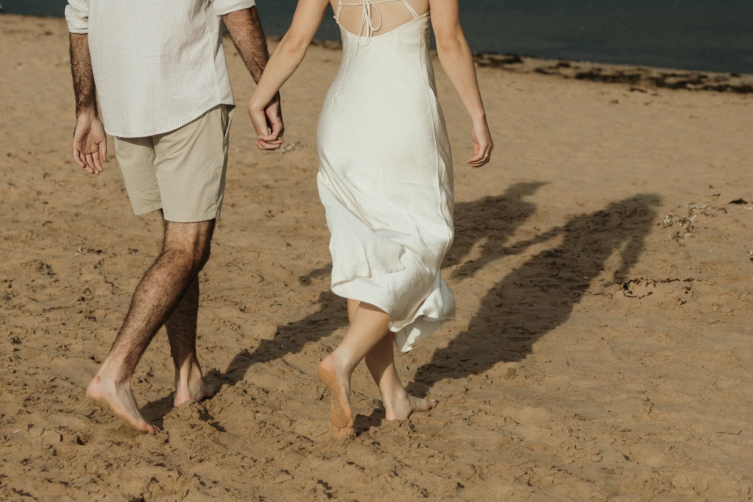 A couple walks barefoot together on sandy beach near lighthouse.