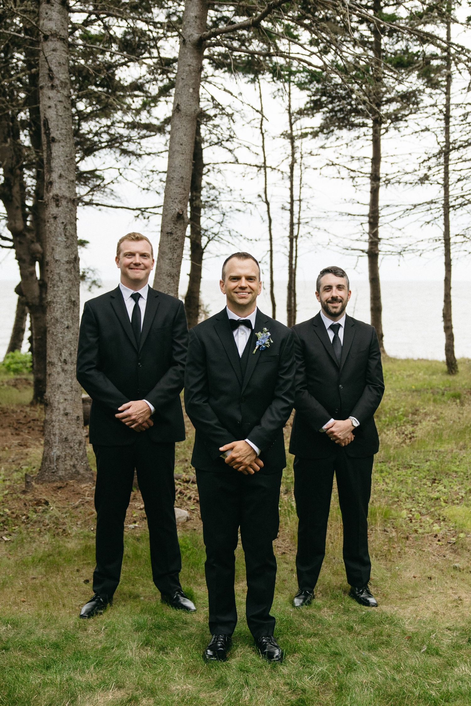 Groom and two groomsmen in black tuxedos posing among tall pine trees near waterfront.