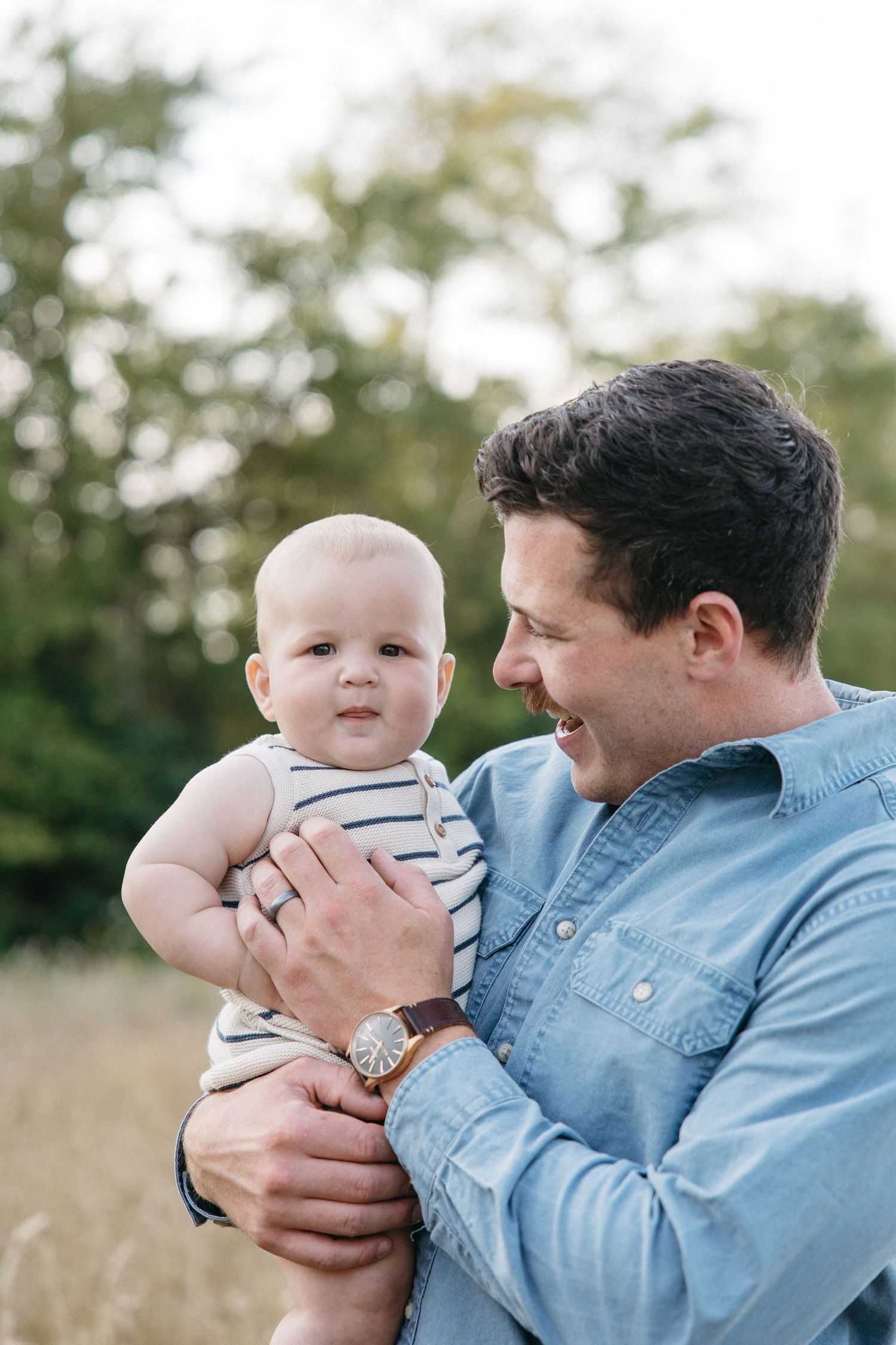 Happy father in denim shirt holds adorable baby in striped onesie outdoors in field.