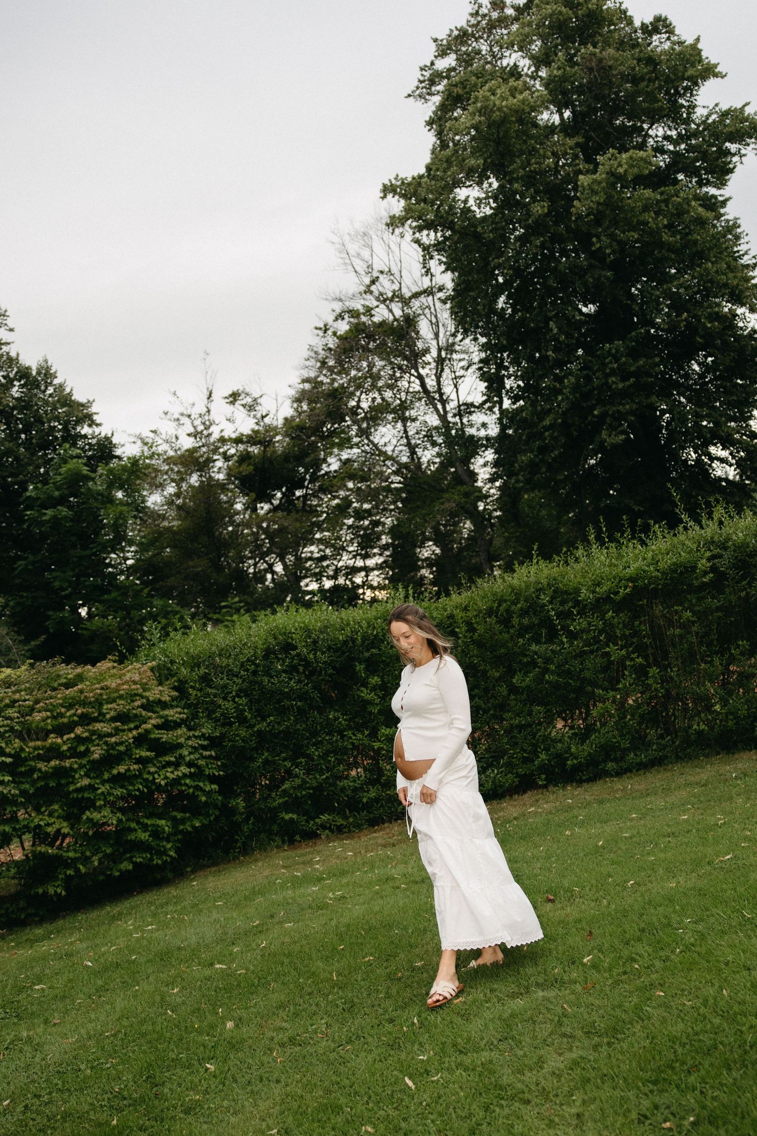 Pregnant woman in white dress walking on green grass in a lush garden.