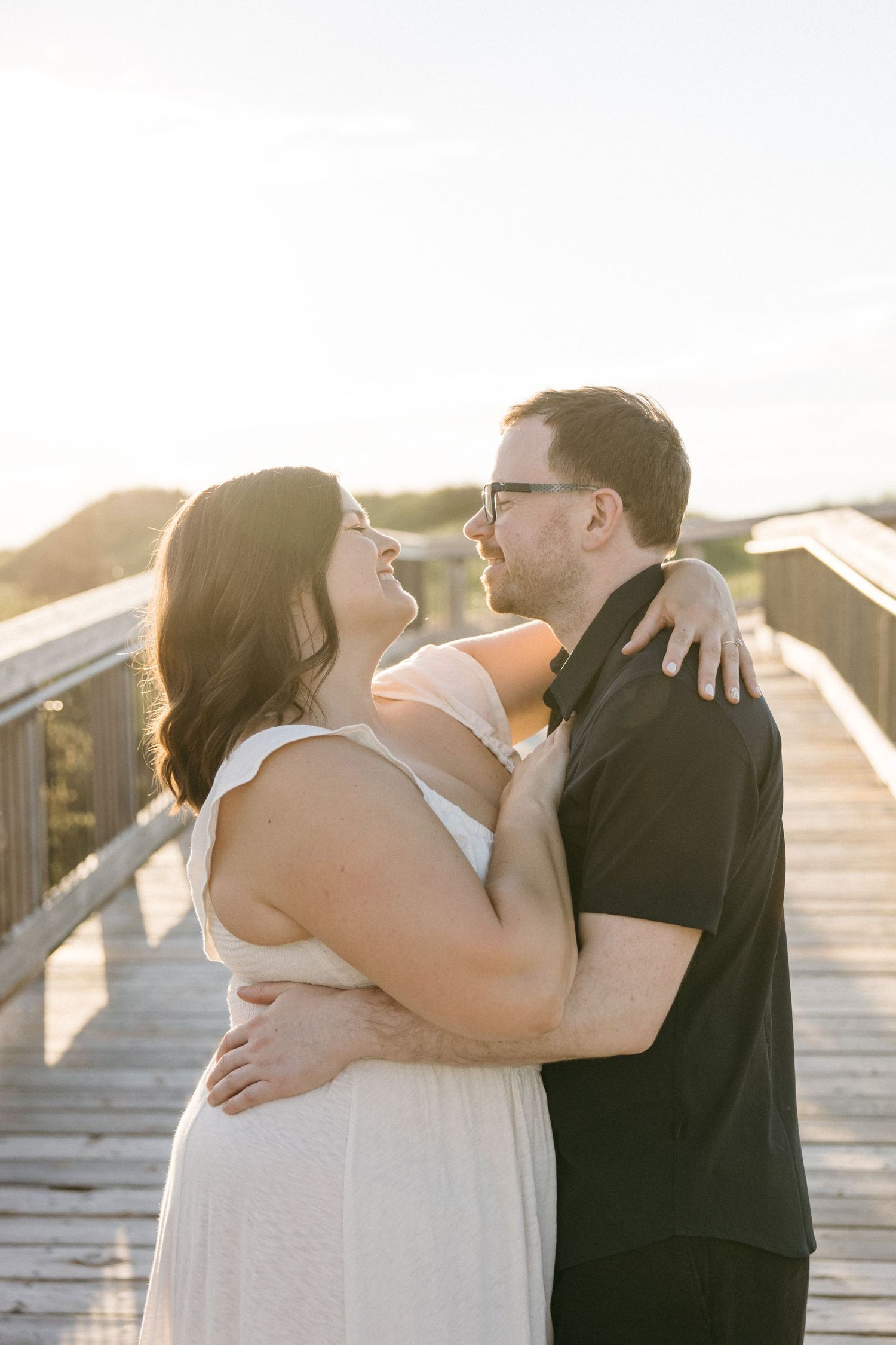 A couple embraces on a wooden boardwalk at golden hour, smiling at each other in a tender romantic moment.
