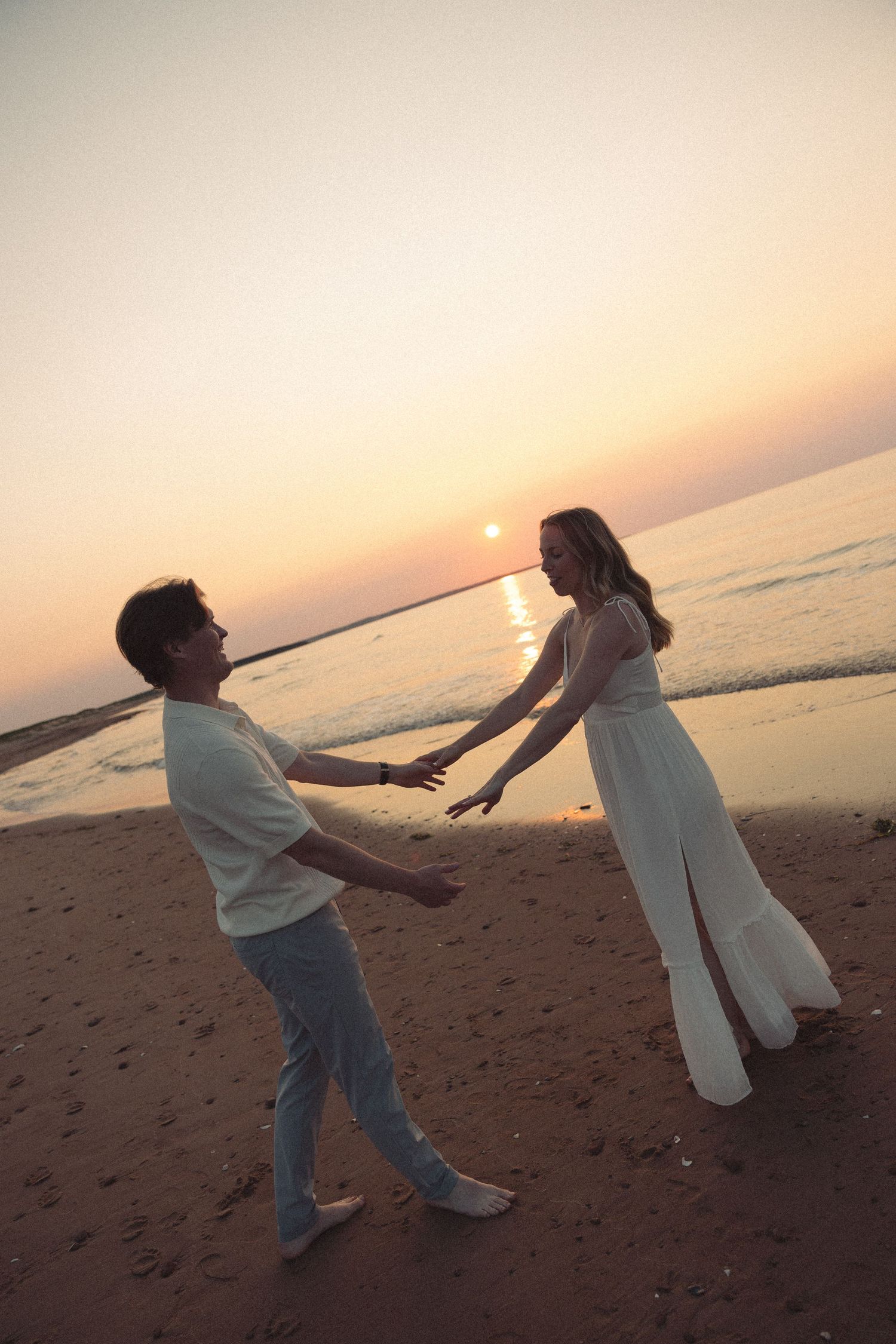 Couple reaching toward each other on sandy beach with vibrant orange sunset over the ocean.